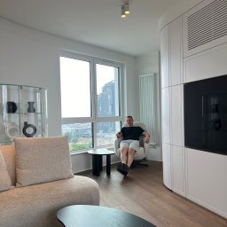 BluMeble - Jasne wnętrze salonu z drewnianą podłogą, beżową sofą i zabudową meblową na ścianie. Mężczyzna siedzi w fotelu na tle okna z widokiem na miasto.