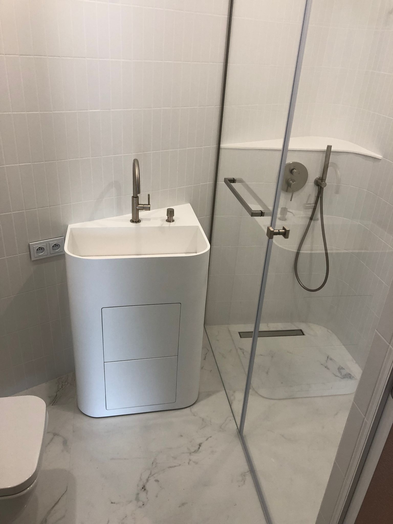 Nowoczesna łazienka z białą, minimalistyczną umywalką z szafką, kabiną prysznicową ze szklaną ścianą i marmurową podłogą. Widoczne detale armatury i płytek.