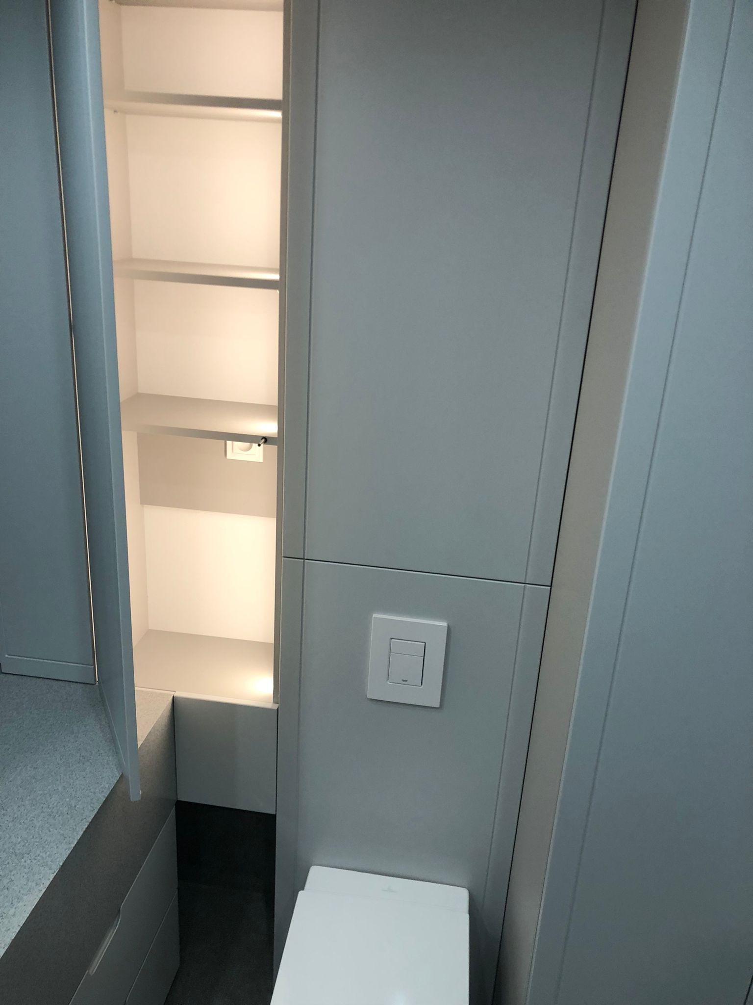 Szara szafa wnękowa z oświetleniem wewnętrznym, otwarte drzwi ukazują puste półki. Widoczny włącznik światła i fragment toalety.