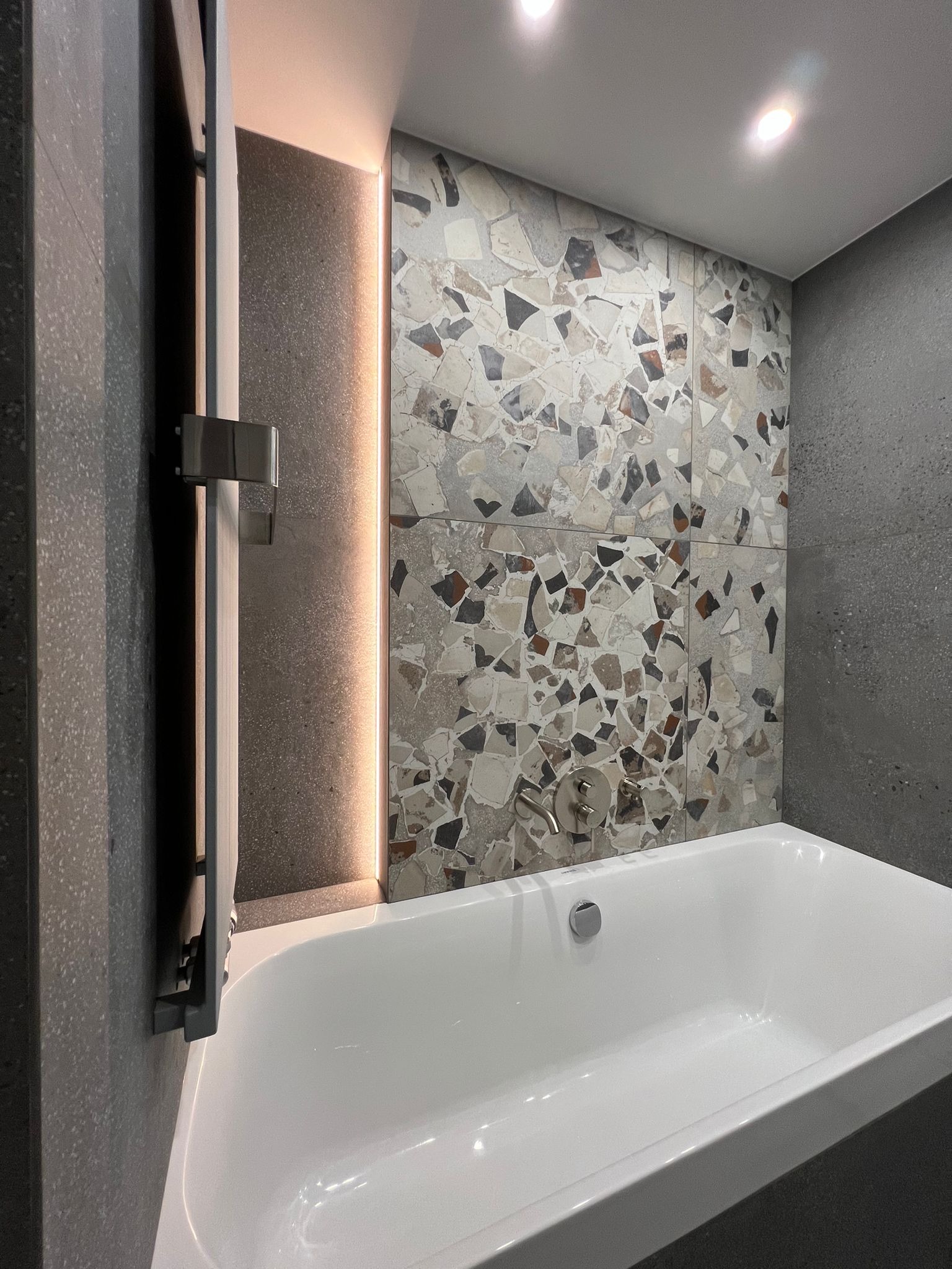 Nowoczesna łazienka z białą wanną, mozaikową ścianą i podświetlanym panelem. Minimalistyczny design, szare płytki, punktowe oświetlenie.