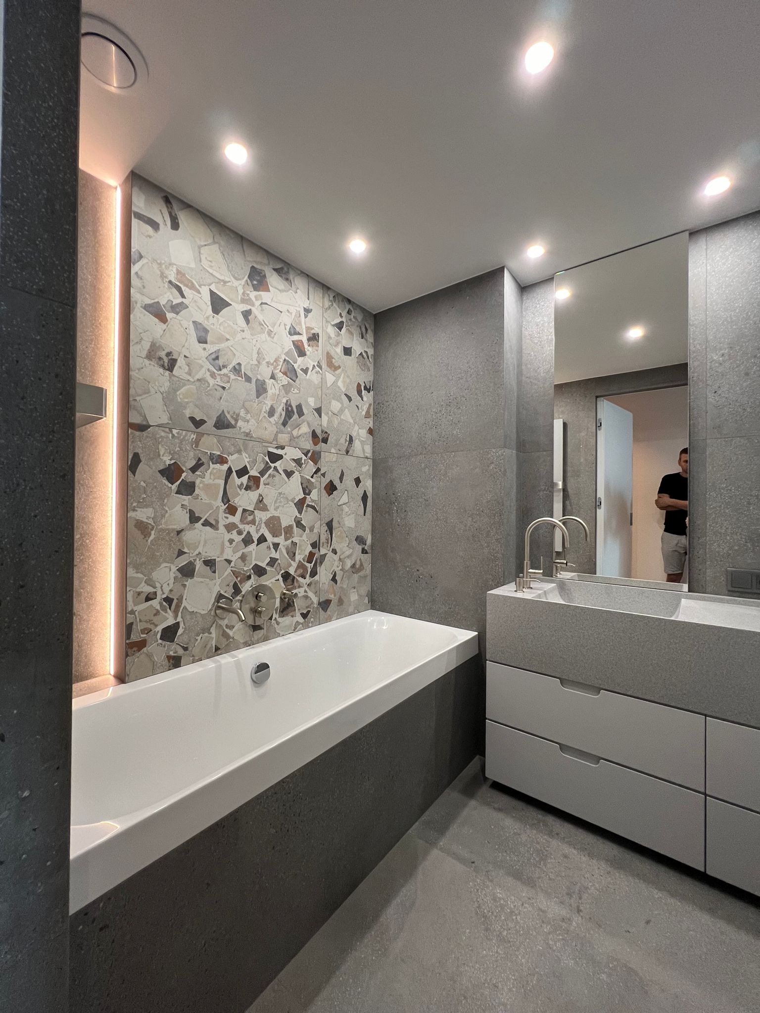 Nowoczesna łazienka z wanną obudowaną szarą mozaiką, umywalką z szafką w odcieniach szarości i dużym lustrem odbijającym fragment wnętrza.