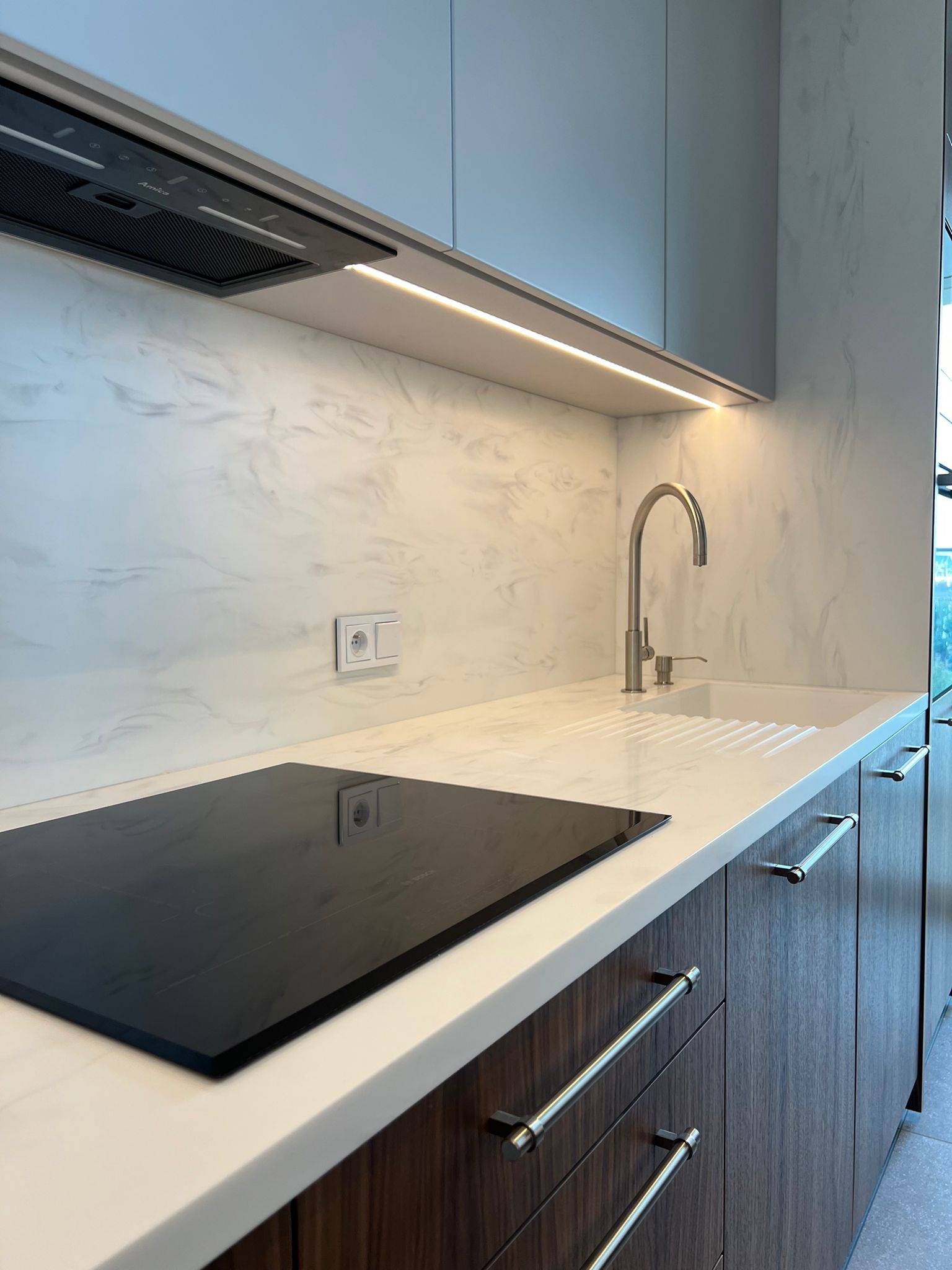 Kompaktowa kuchnia: płyta indukcyjna, bateria zlewozmywakowa, marmurowy panel ścienny i oświetlenie LED pod szafkami. Fronty szafek w odcieniach brązu i szarości.