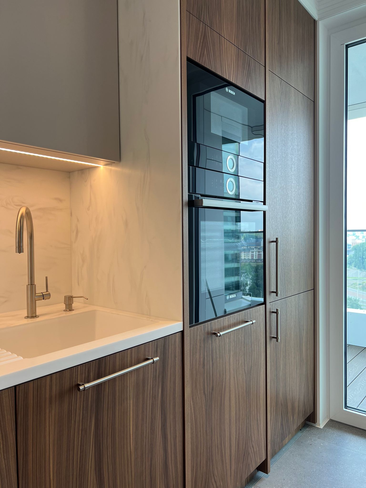 Nowoczesna zabudowa kuchenna w Warszawie: fornir orzech włoski, sprzęt Bosch, biały blat i zlew, minimalistyczna armatura, oświetlenie LED pod szafkami.