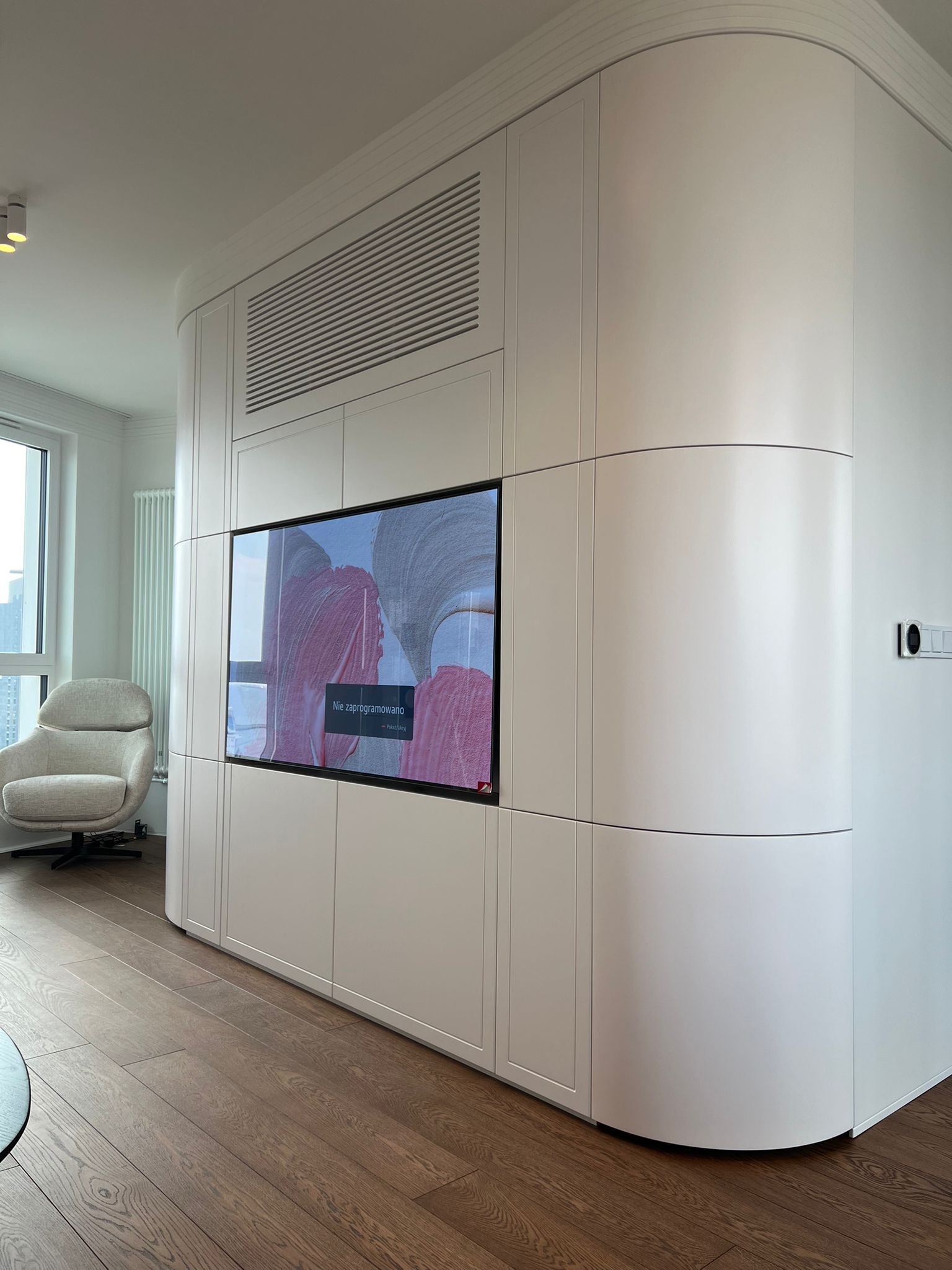 Biała zabudowa meblowa z zaokrąglonym narożnikiem, wkomponowany telewizor, drewniana podłoga, fotel w tle, nowoczesny design.