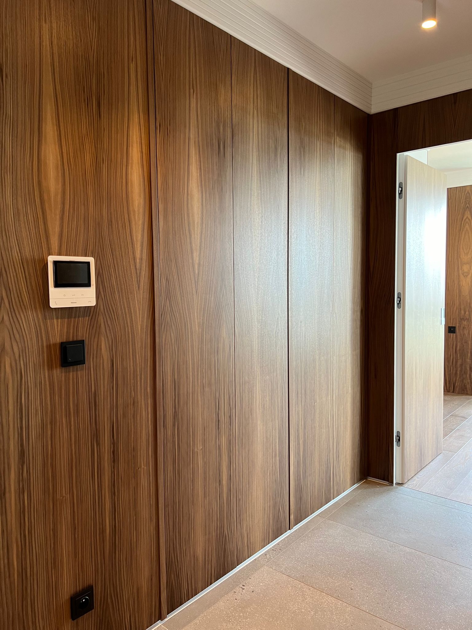 Ściana z forniru orzechowego z ukrytymi drzwiami i szafami, minimalistyczny design, biała listwa przypodłogowa i sufitowa, wideodomofon na ścianie.