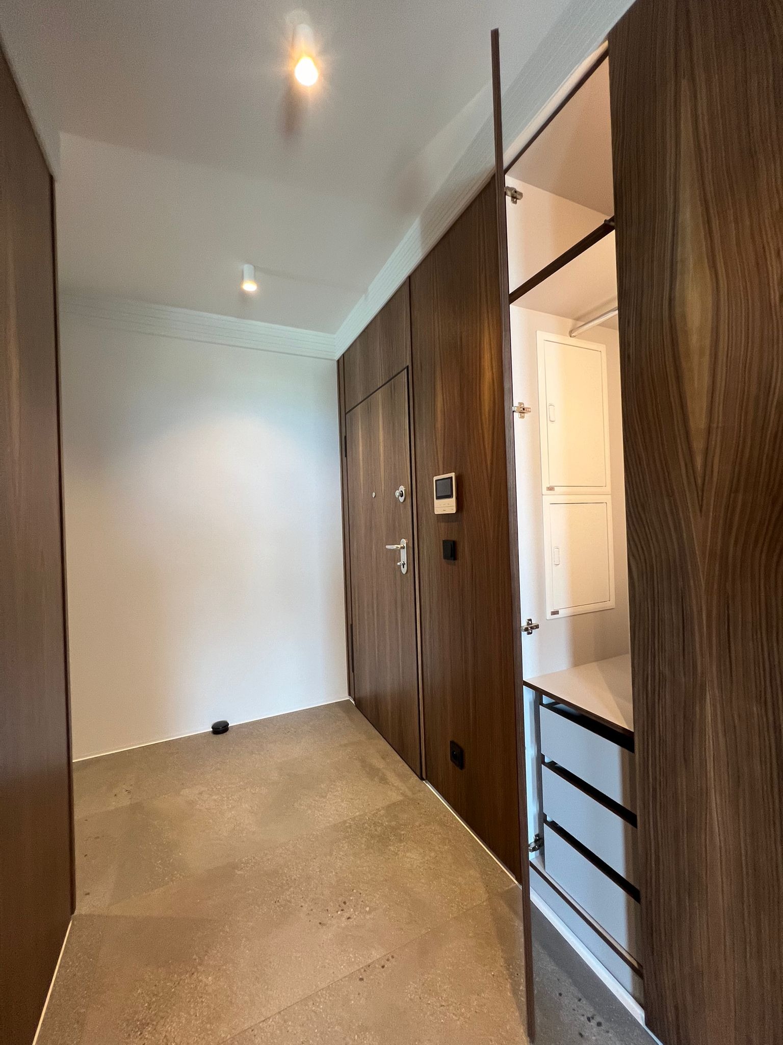 Hol wejściowy z zabudową szafy z forniru orzechowego, częściowo otwarty, ukazujący szuflady i białe drzwiczki rewizyjne. Minimalistyczny design, ciepłe oświetlenie.