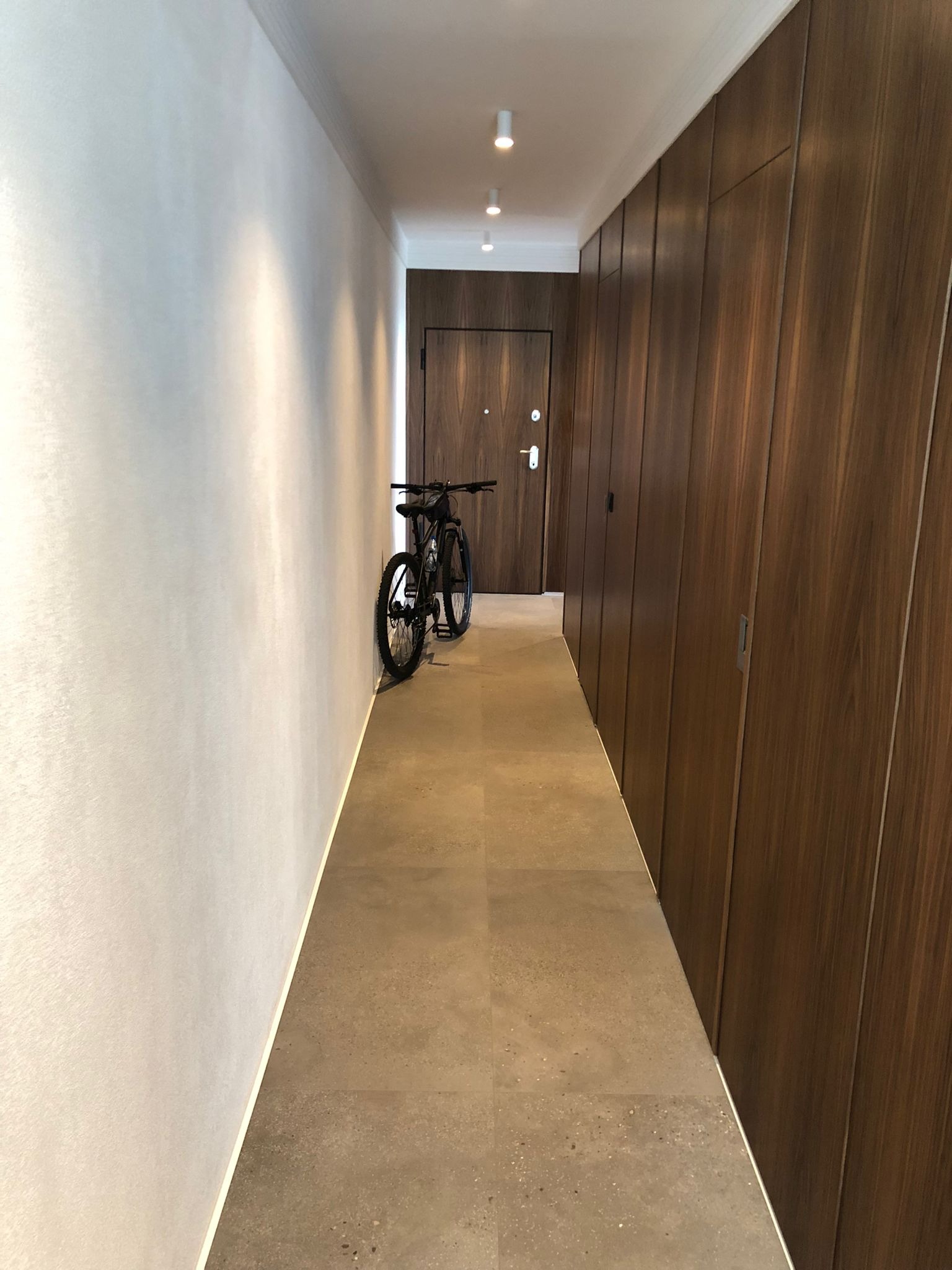 Długi korytarz z gładkimi, białymi ścianami i zabudową z ciemnego drewna. Na podłodze płytki w odcieniach beżu. Rower oparty o ścianę. Minimalistyczne oświetlenie sufitowe.