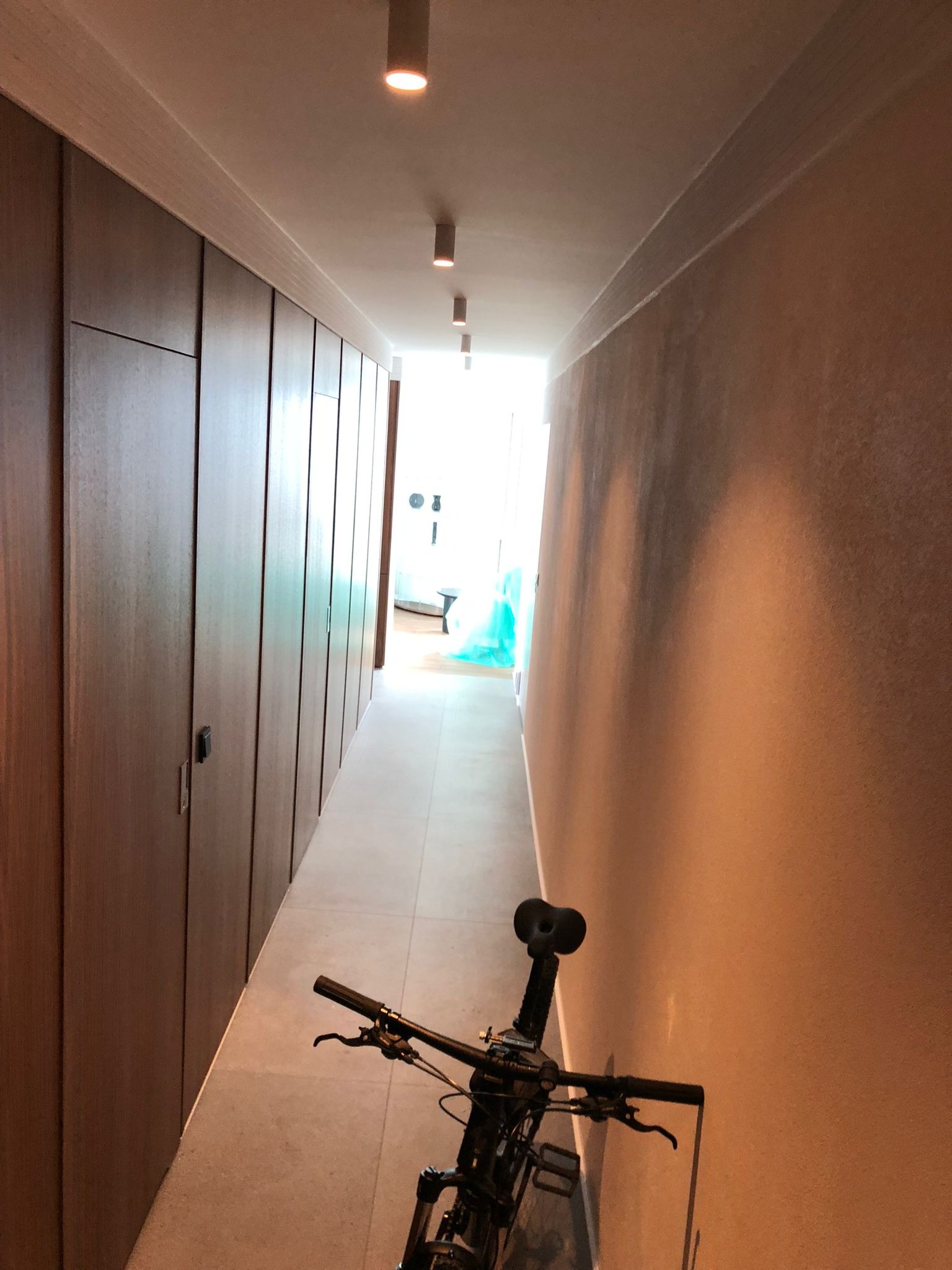 Długi korytarz z zabudową szaf w odcieniach brązu i beżowymi ścianami. W tle widoczne jasne pomieszczenie. Na pierwszym planie fragment roweru.