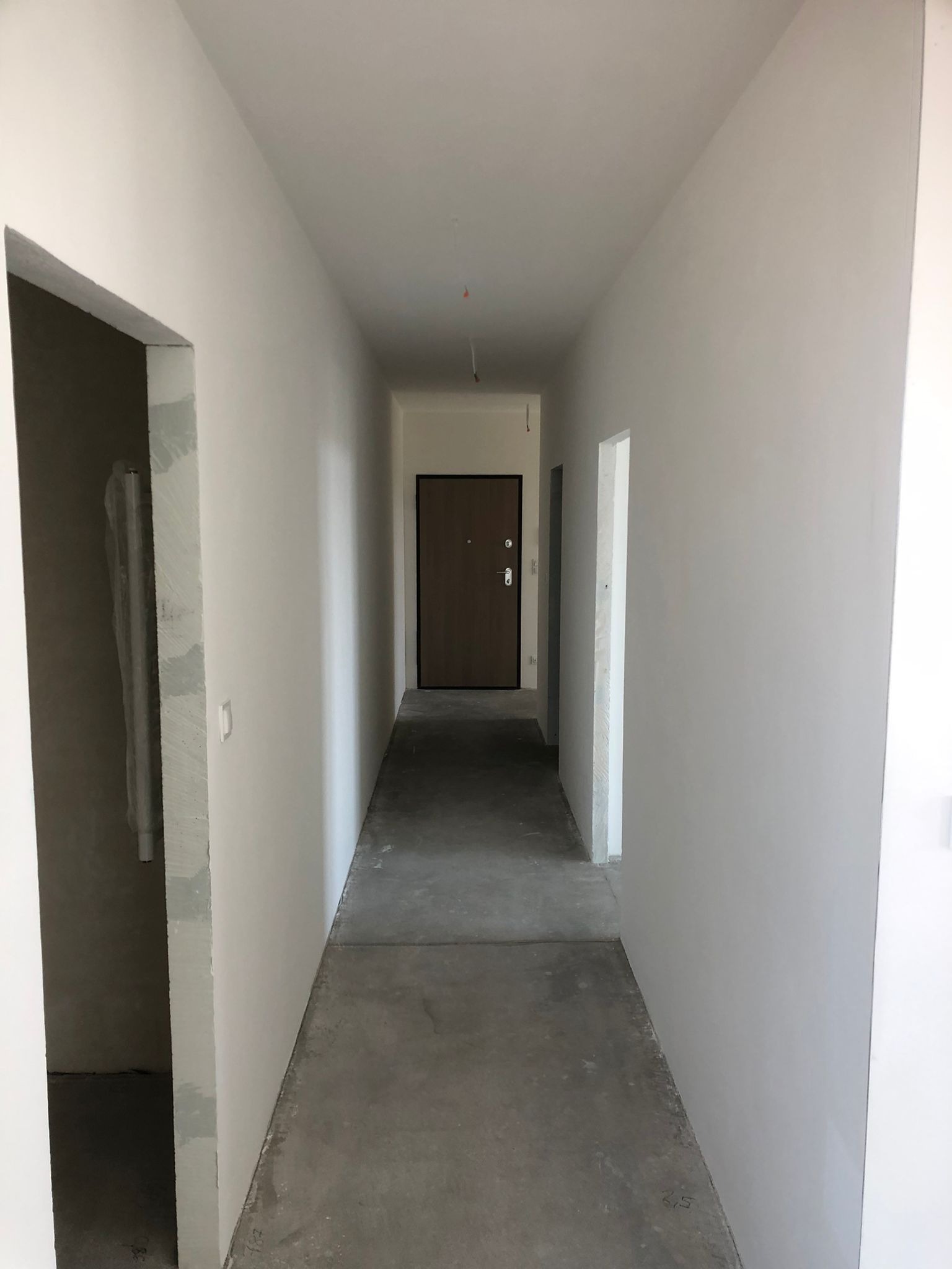 Korytarz w trakcie wykończenia: świeżo pomalowane ściany w odcieniach bieli, surowa betonowa podłoga. Widoczne otwory drzwiowe i drzwi wejściowe w głębi.