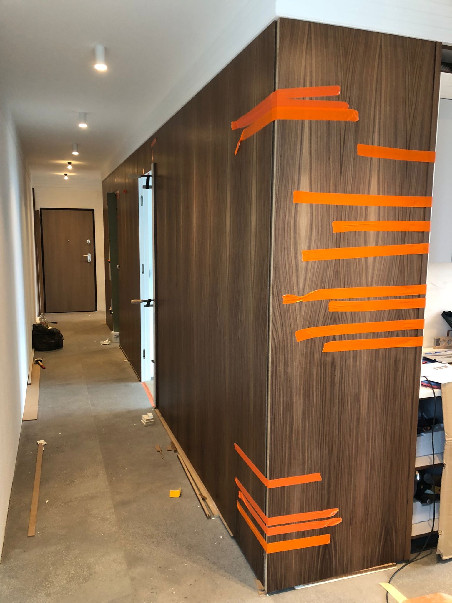 Montaż szafy wnękowej z okleiną drewnopodobną, zabezpieczoną pomarańczową taśmą. Widoczny fragment korytarza z drzwiami i oświetleniem.