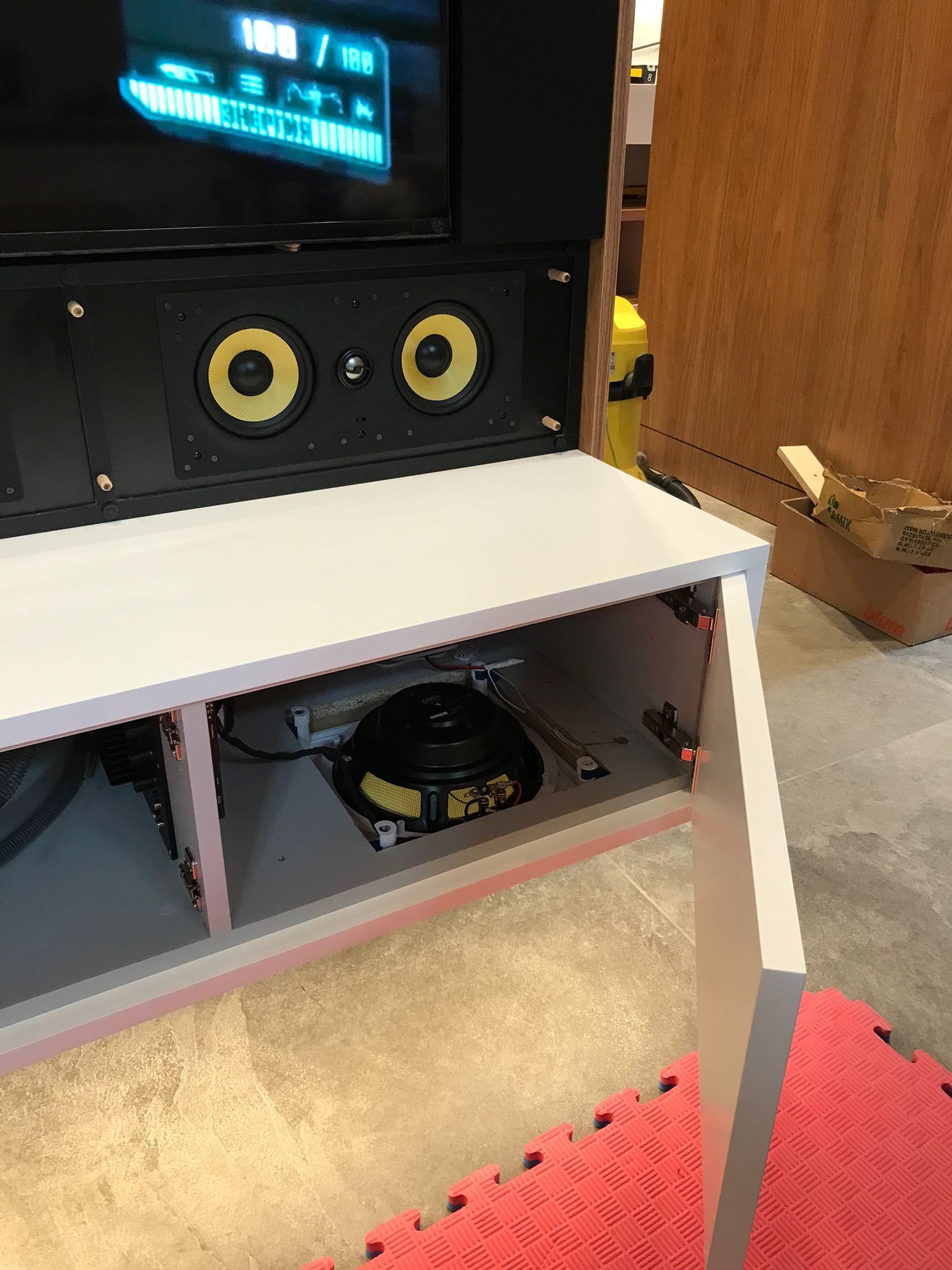 Biała szafka RTV z otwartymi drzwiczkami, ukazująca głośnik basowy wewnątrz. Nad szafką widoczny ekran i głośniki. Minimalistyczny design.