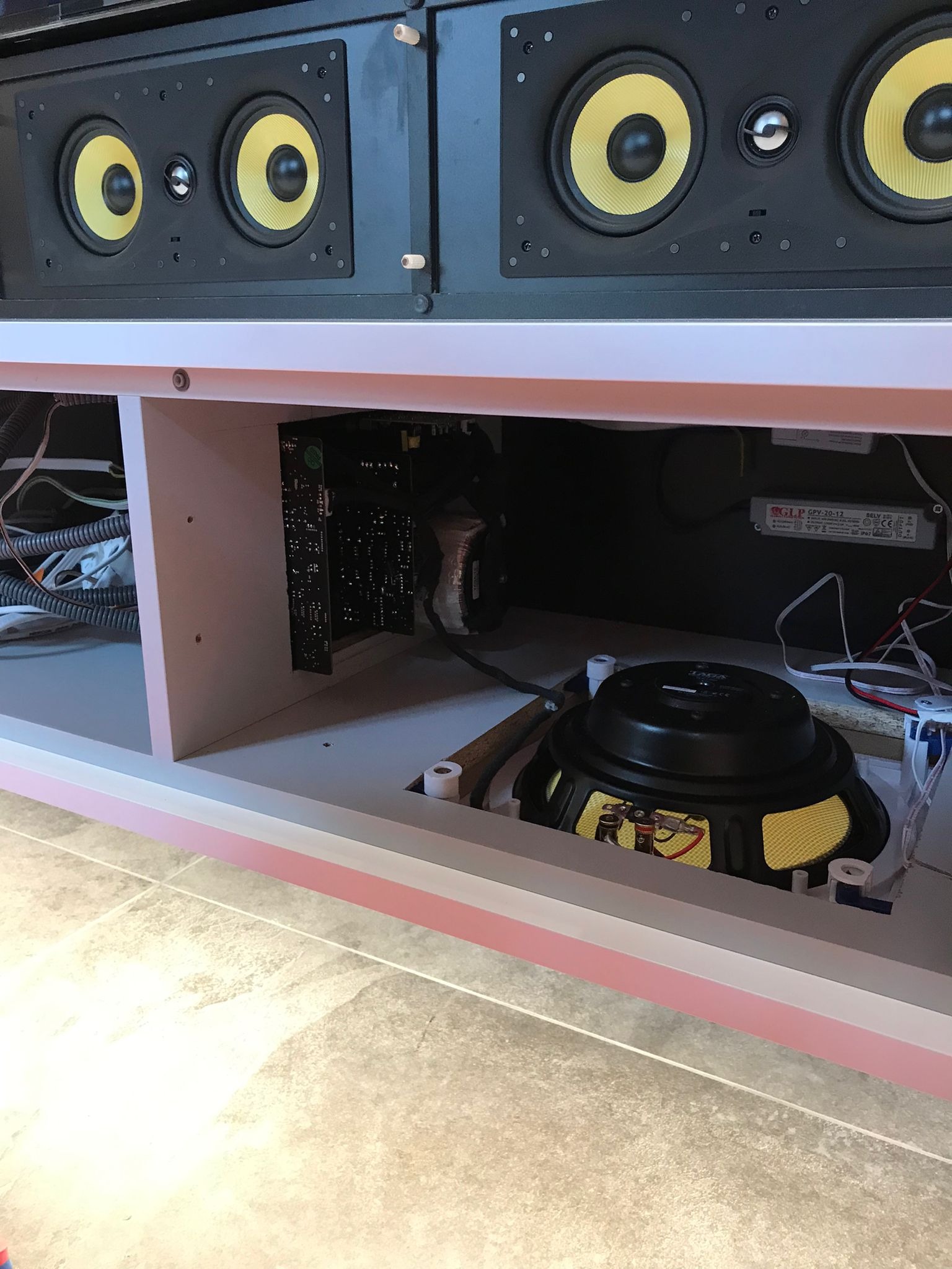 Wnętrze szafki RTV z widocznymi głośnikami z żółtymi membranami, elektroniką i okablowaniem. Widok z dołu, ukazujący instalację sprzętu audio.