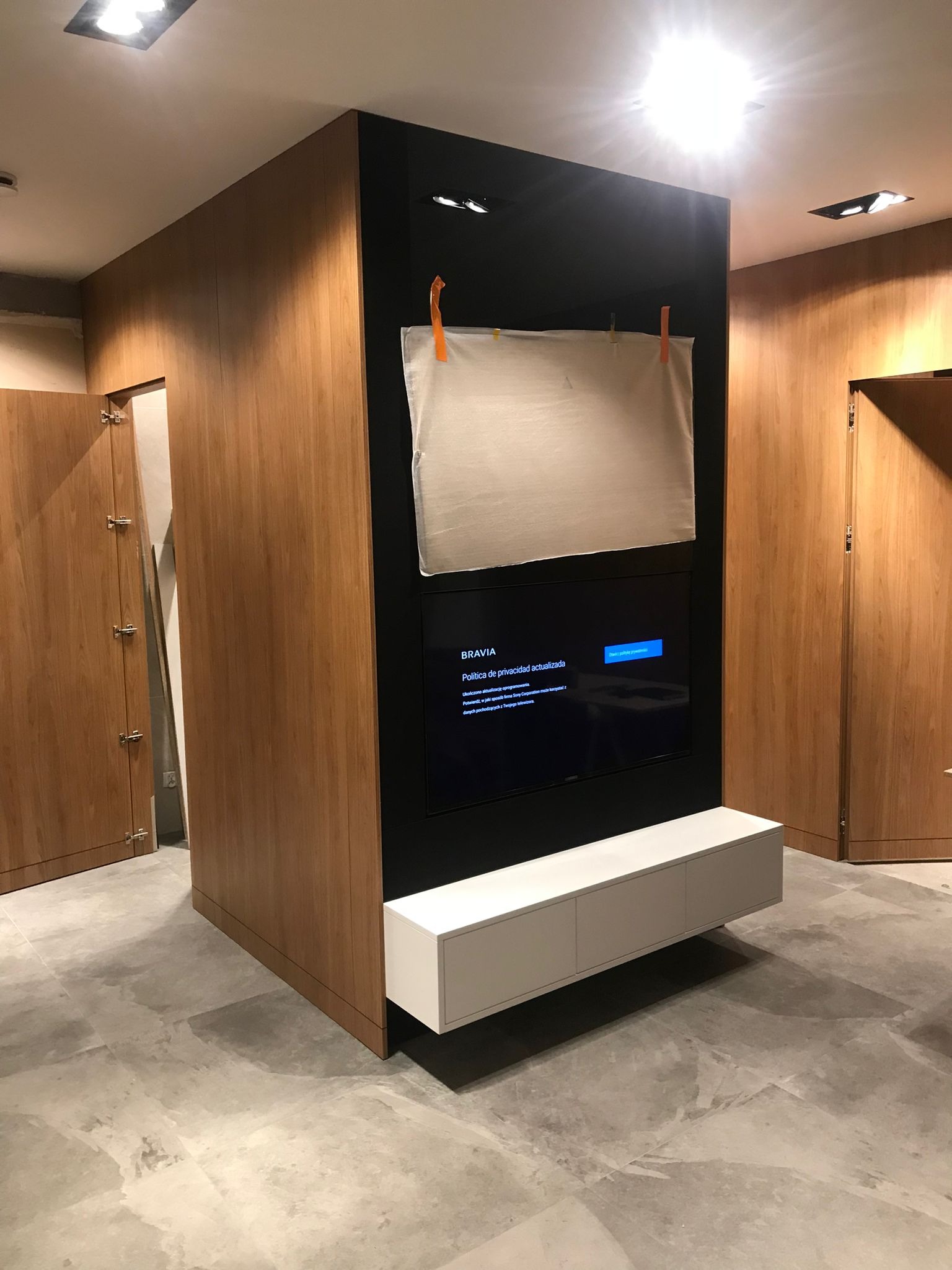 Nowoczesna zabudowa RTV z drewnianymi frontami i czarnym panelem. Podwieszana szafka RTV w kolorze białym. Białe płótno zawieszone na klipsach.