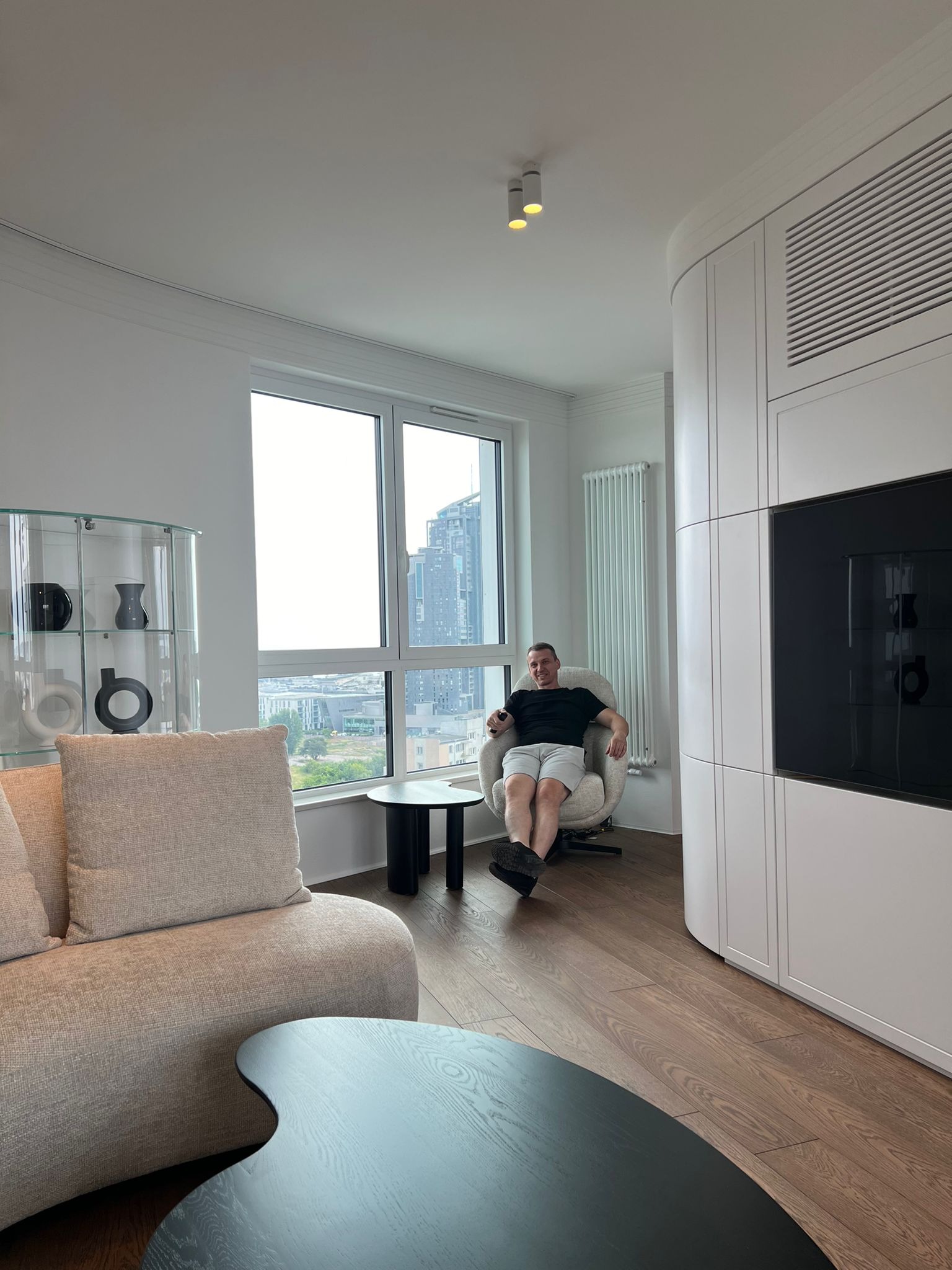 Jasne wnętrze salonu z drewnianą podłogą, beżową sofą i zabudową meblową na ścianie. Mężczyzna siedzi w fotelu na tle okna z widokiem na miasto.