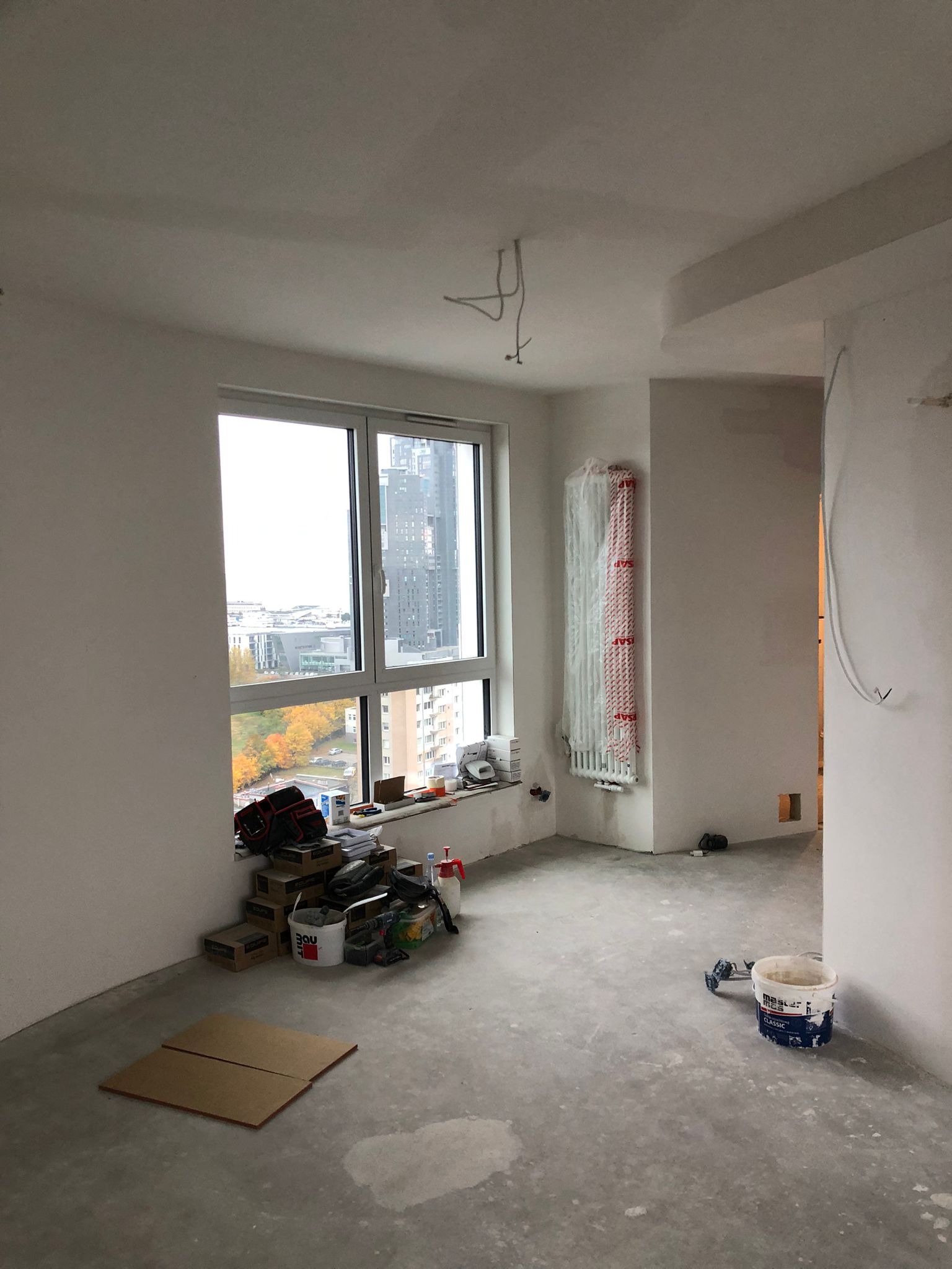 Wnętrze pokoju w trakcie remontu: ściany po gładziowaniu, okno z widokiem na miasto, narzędzia i materiały budowlane na podłodze. Grzejnik zabezpieczony folią.