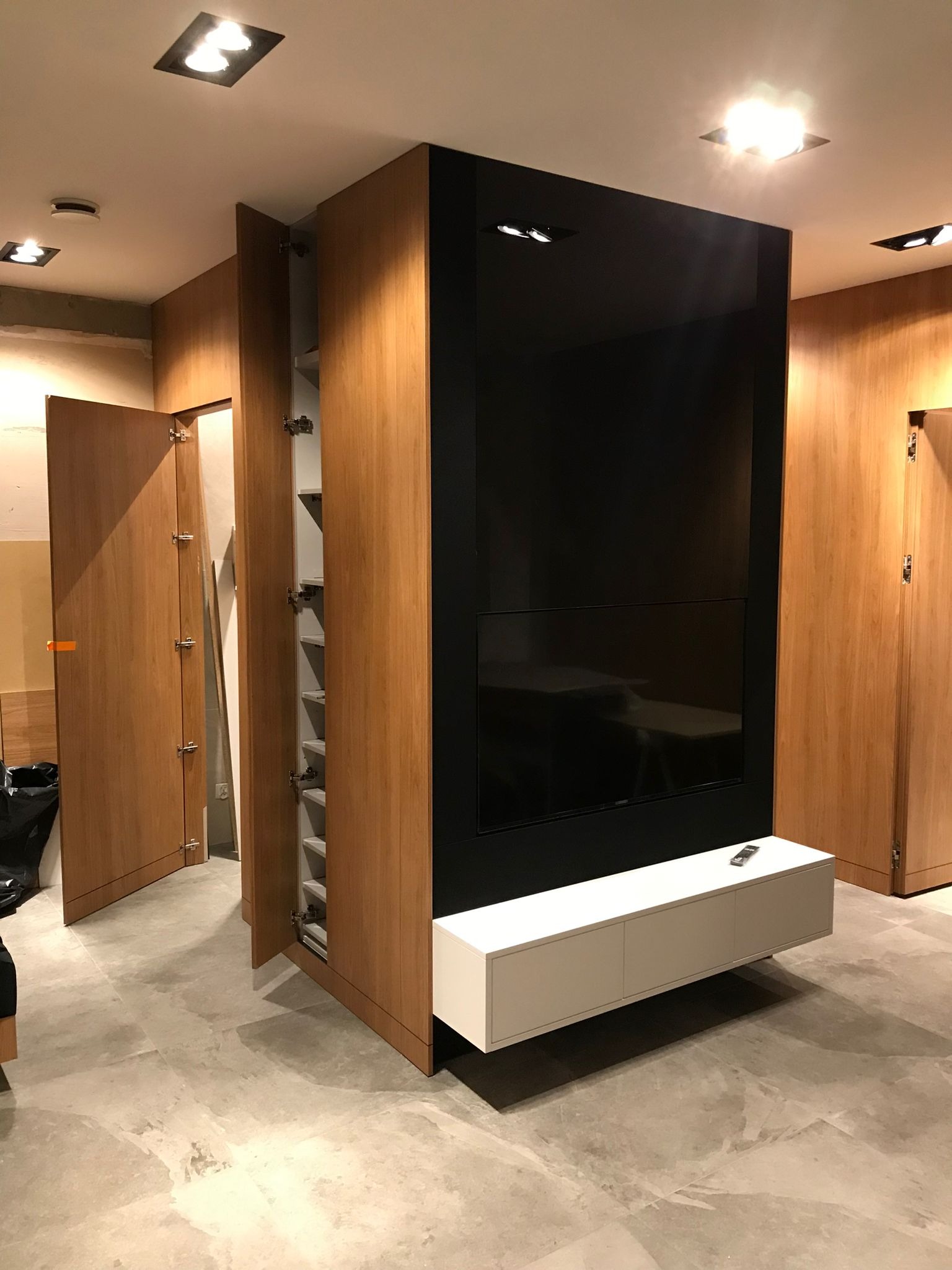 Nowoczesna zabudowa meblowa z drewnianymi szafami, czarnym panelem TV i białą szafką RTV. Otwarta szafa z półkami. Minimalistyczny design, sufit z oświetleniem.