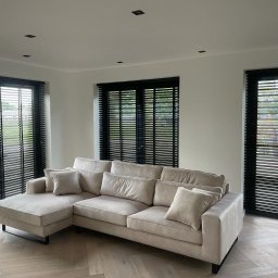 ALVECO Paweł Kępczyński - Salon z beżową sofą i trzema oknami z żaluzjami w czarnych ramach. Parkiet w jodełkę i minimalistyczne oświetlenie sufitowe.