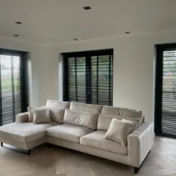 Salon z beżową sofą i trzema oknami z żaluzjami w czarnych ramach. Parkiet w jodełkę i minimalistyczne oświetlenie sufitowe.