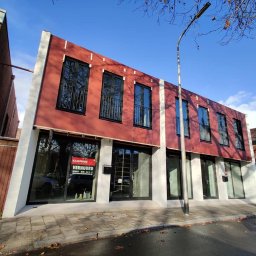 ALVECO Paweł Kępczyński - Nowoczesny budynek z czerwono-białą fasadą i czarnymi oknami, widziany z ulicy w słoneczny dzień. Widoczne okna na piętrze z ozdobnymi balustradami.