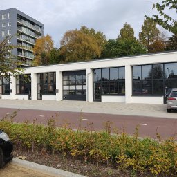 ALVECO Paweł Kępczyński - Biały budynek z czarnymi, przeszklonymi segmentowymi drzwiami garażowymi i oknami, widok z zewnątrz, w tle drzewa i budynek mieszkalny.