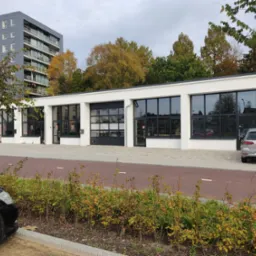 Biały budynek z czarnymi, przeszklonymi segmentowymi drzwiami garażowymi i oknami, widok z zewnątrz, w tle drzewa i budynek mieszkalny.