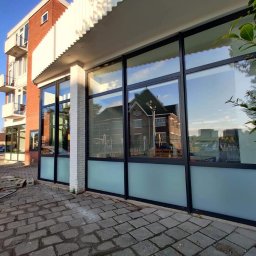 ALVECO Paweł Kępczyński - Elewacja budynku z dużymi oknami z czarnymi ramami i mleczną szybą u dołu, odbijającymi otoczenie. Widok z zewnątrz na chodnik z kostki brukowej.