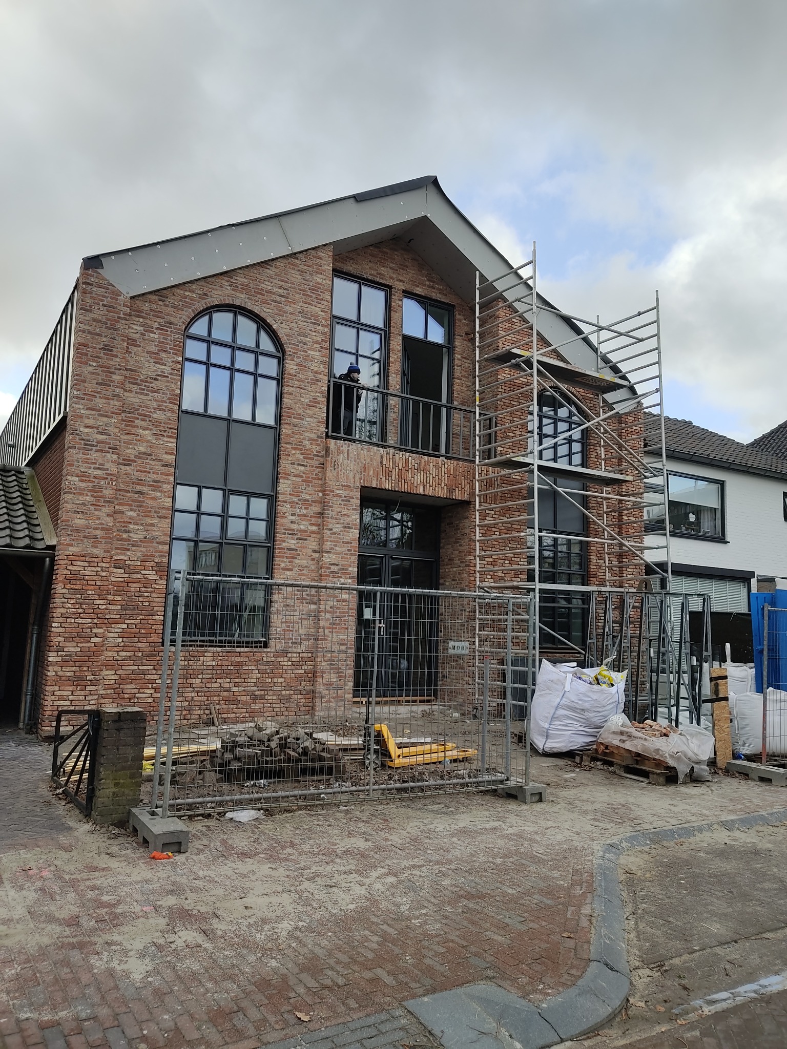 Ceglany dom w budowie z czarnymi oknami i rusztowaniem. Widoczne materiały budowlane i ogrodzenie tymczasowe. Osoba na balkonie. Pochmurne niebo w tle.