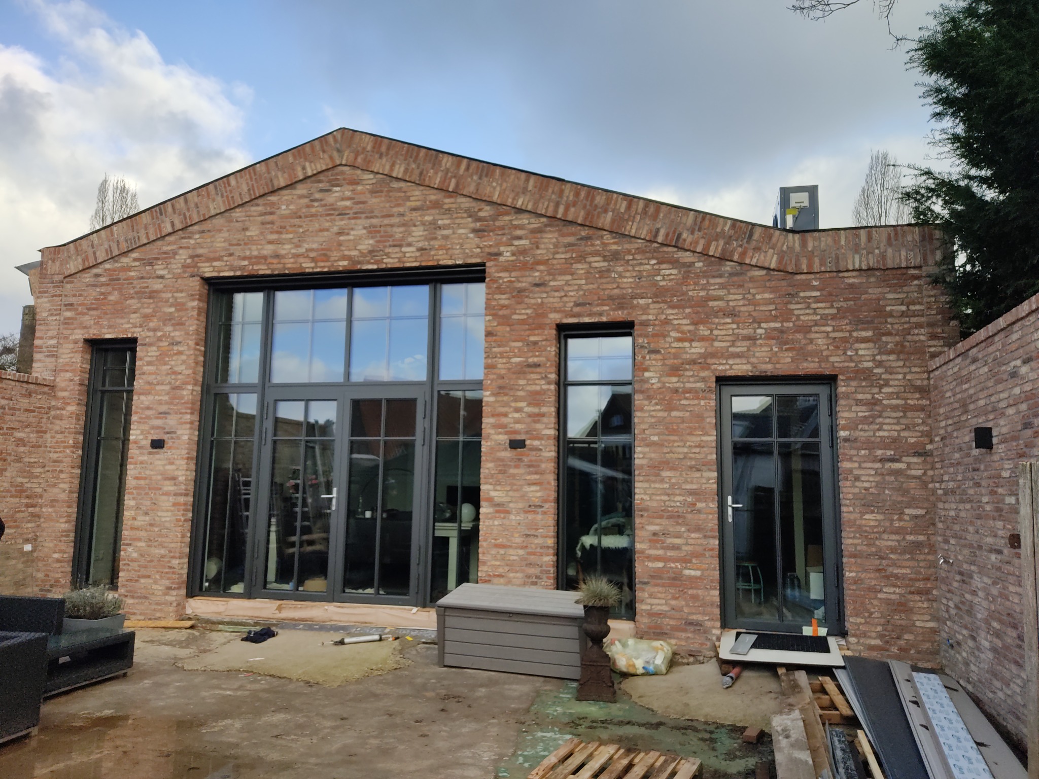 Nowoczesny dom z cegły z dużymi, czarnymi oknami i drzwiami. Widoczne elementy montażowe i palety, sugerujące świeżą instalację.