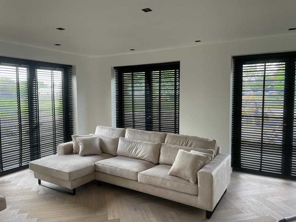 Salon z beżową sofą i trzema oknami z żaluzjami w czarnych ramach. Parkiet w jodełkę i minimalistyczne oświetlenie sufitowe.