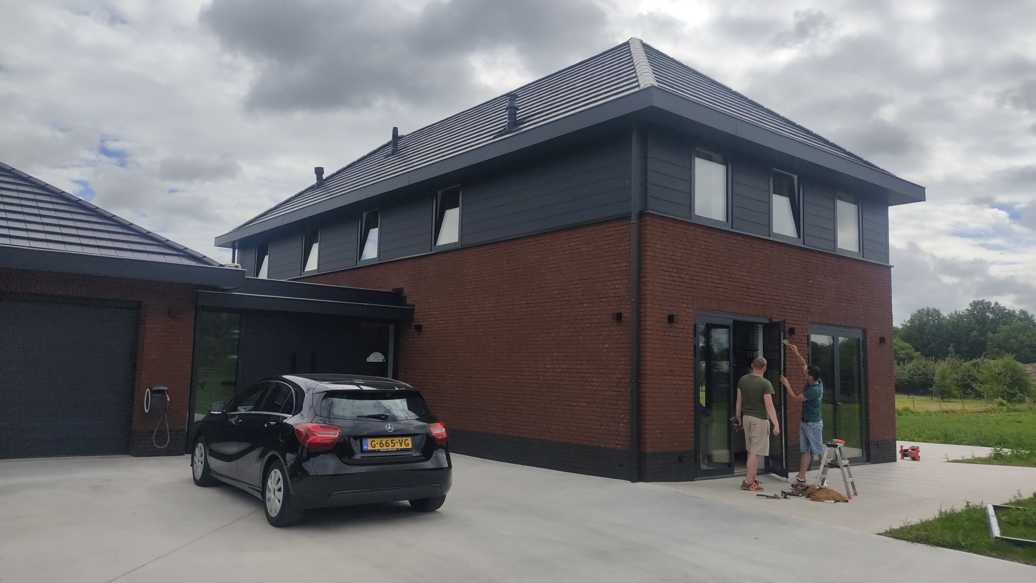 Nowoczesny dom z ciemną stolarką okienną i przesuwnymi drzwiami tarasowymi. Dwóch monterów przy pracy, narzędzia na zewnątrz. Elegancki design.