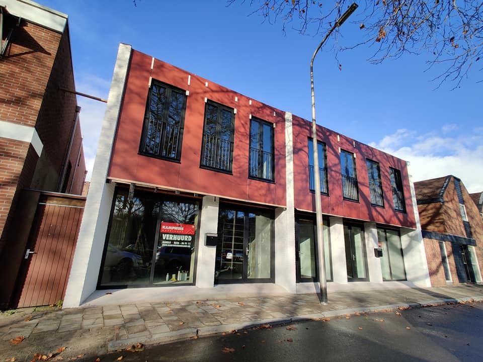 Nowoczesny budynek z czerwono-białą fasadą i czarnymi oknami, widziany z ulicy w słoneczny dzień. Widoczne okna na piętrze z ozdobnymi balustradami.