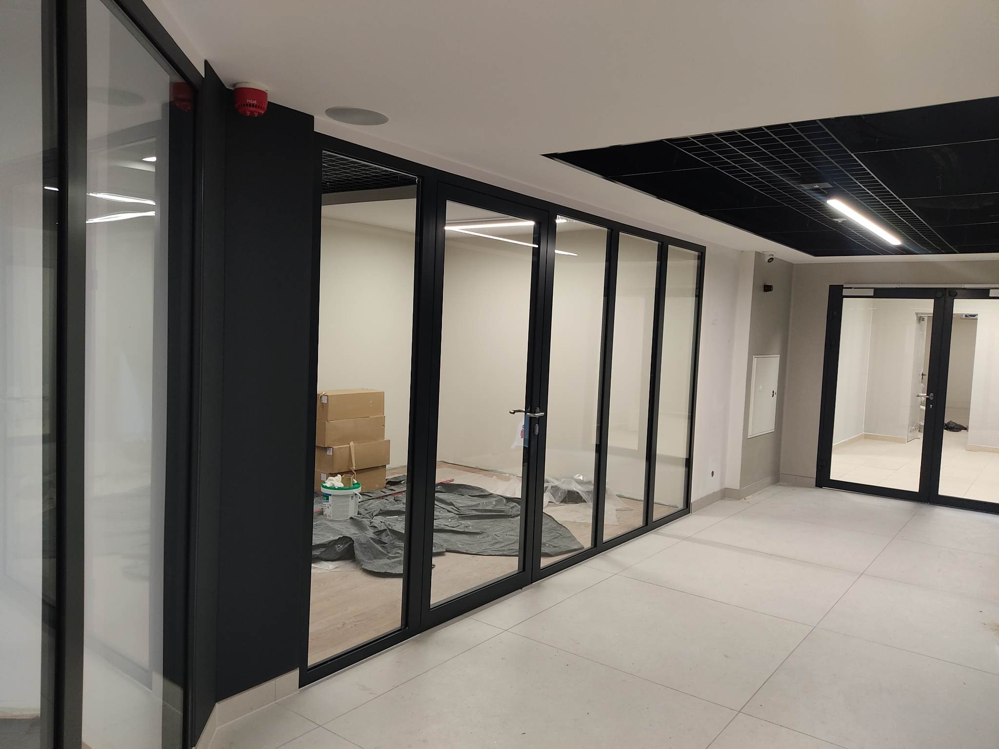 Szklane drzwi przesuwne z czarnymi ramami oddzielają przestrzeń biurową. Widoczne kartony i folia ochronna na podłodze wewnątrz.
