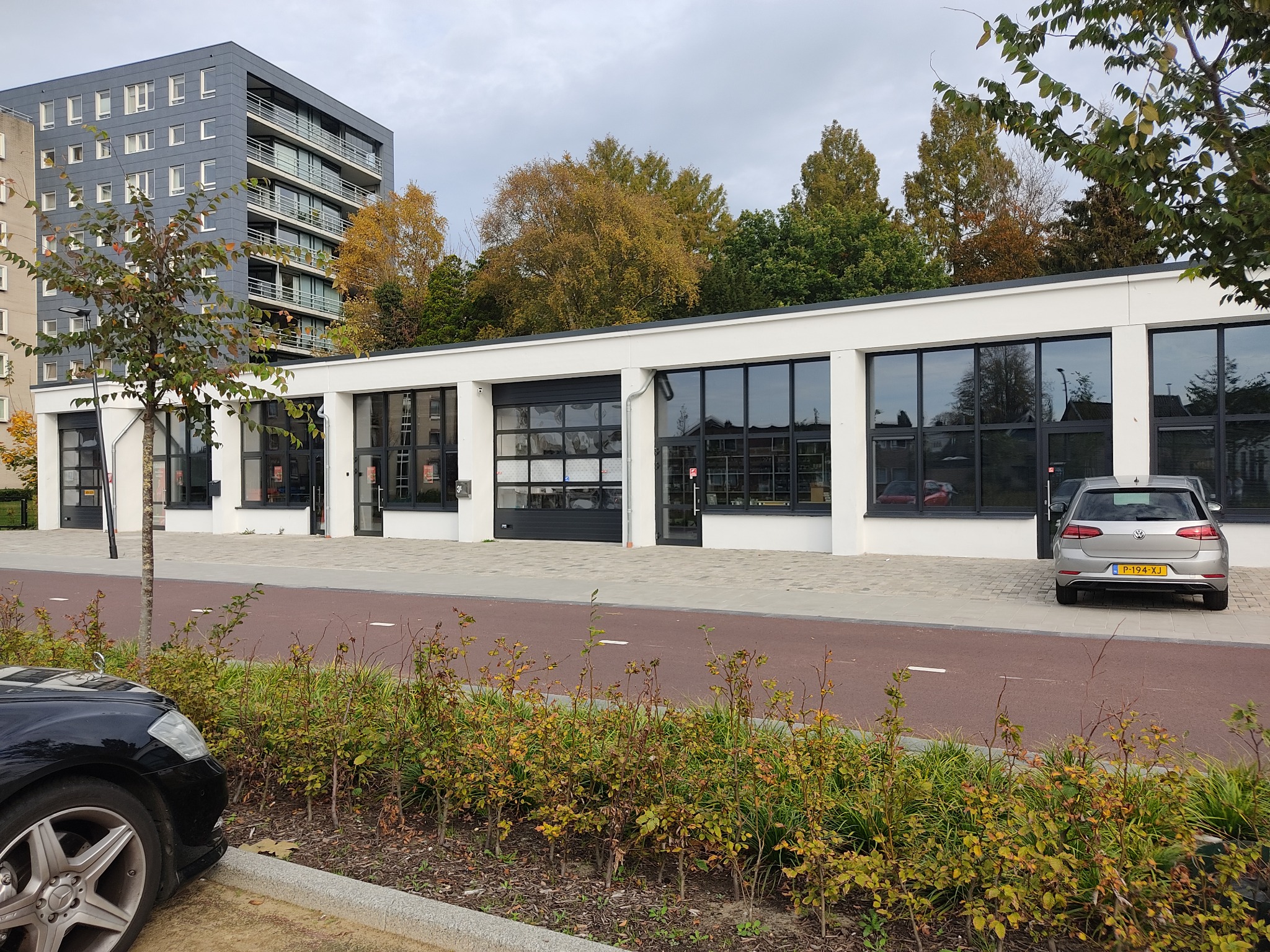 Biały budynek z czarnymi, przeszklonymi segmentowymi drzwiami garażowymi i oknami, widok z zewnątrz, w tle drzewa i budynek mieszkalny.