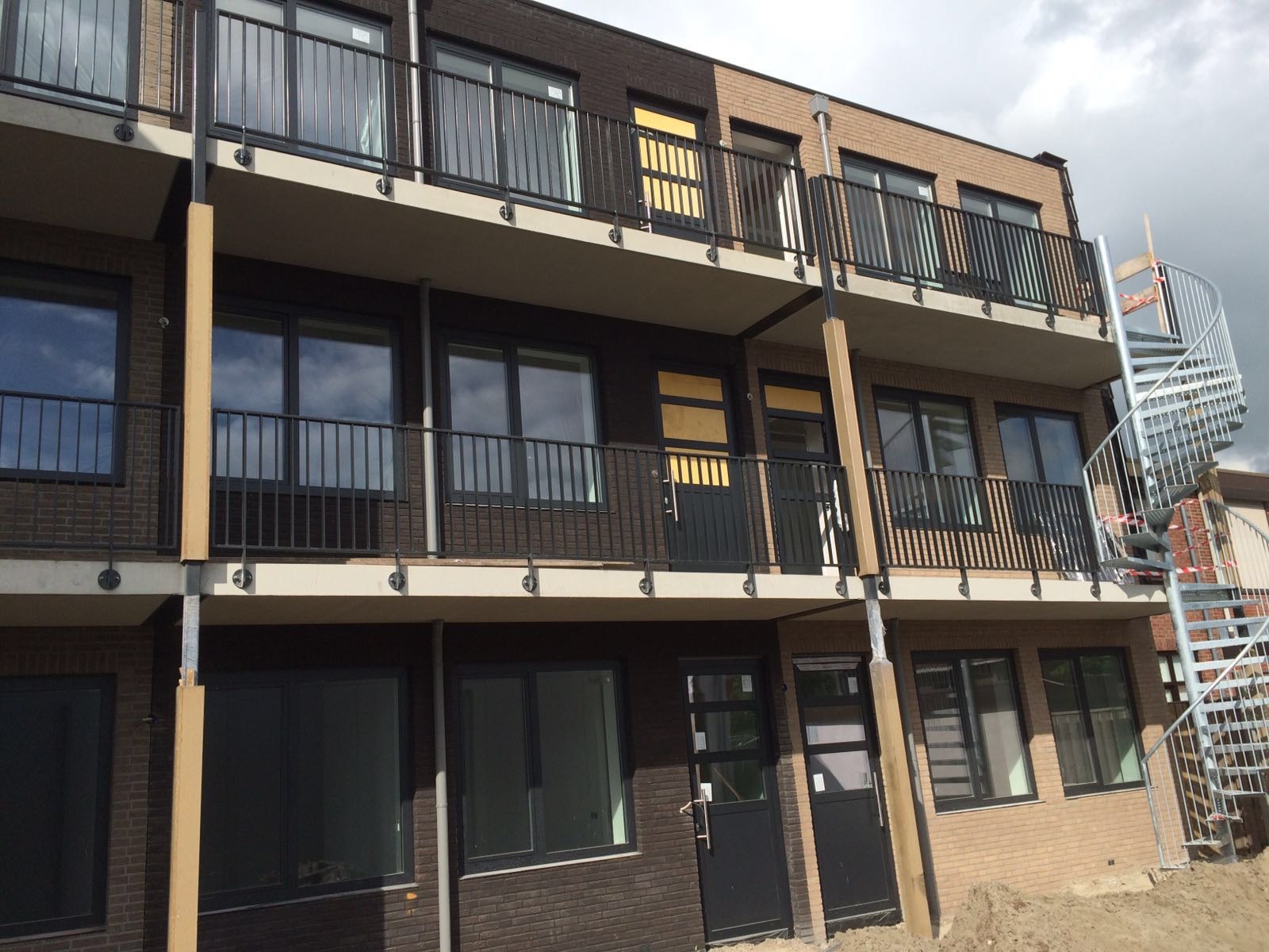 Nowoczesny budynek wielorodzinny z balkonami, czarne okna i drzwi, spiralne schody zewnętrzne, elewacja z cegły w dwóch odcieniach brązu. Częściowo widoczne niebo.