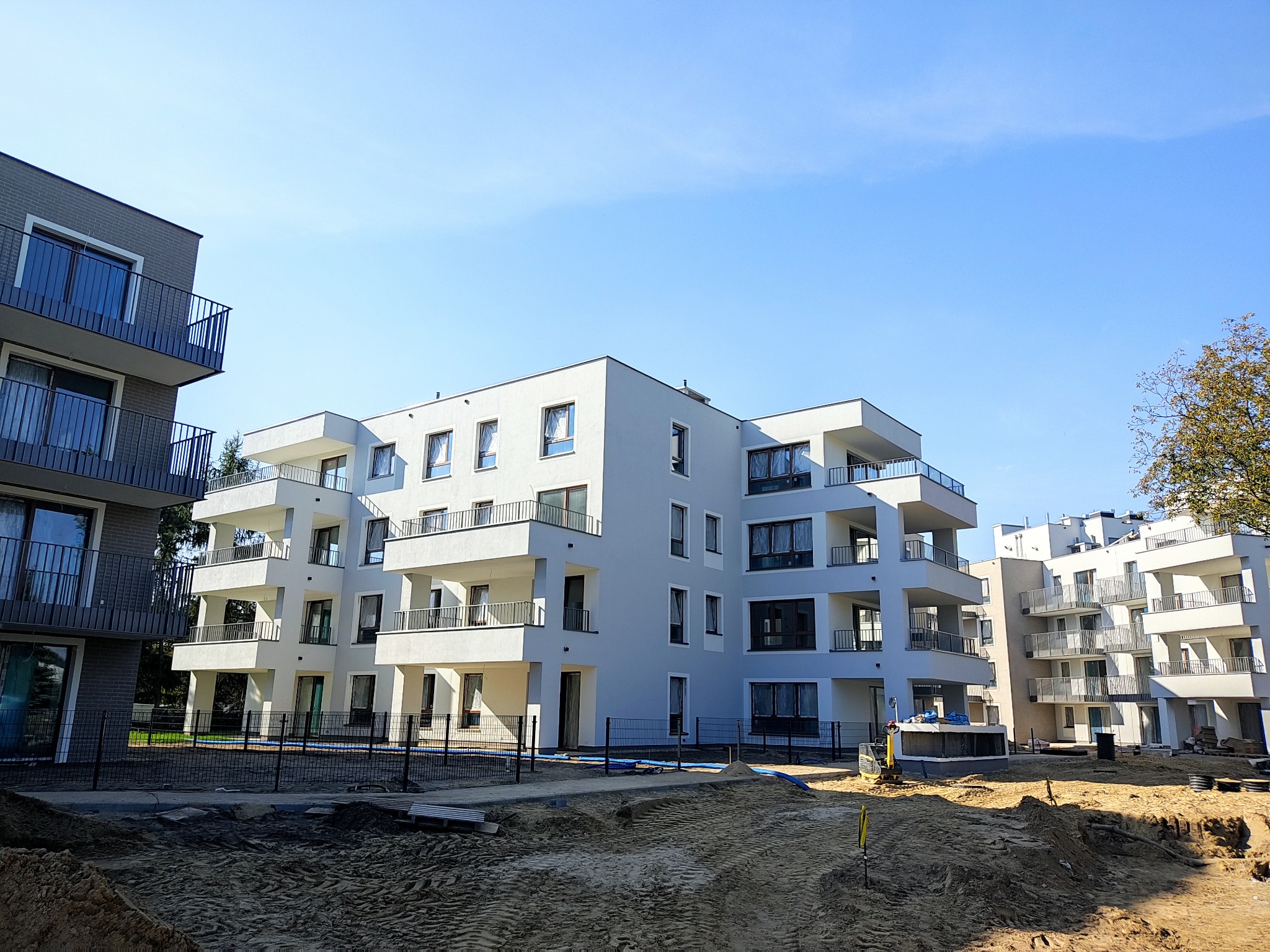 Nowoczesny, trzypiętrowy apartamentowiec z balkonami i szarymi oknami, widziany z poziomu gruntu na placu budowy w słoneczny dzień. Minimalistyczna architektura.