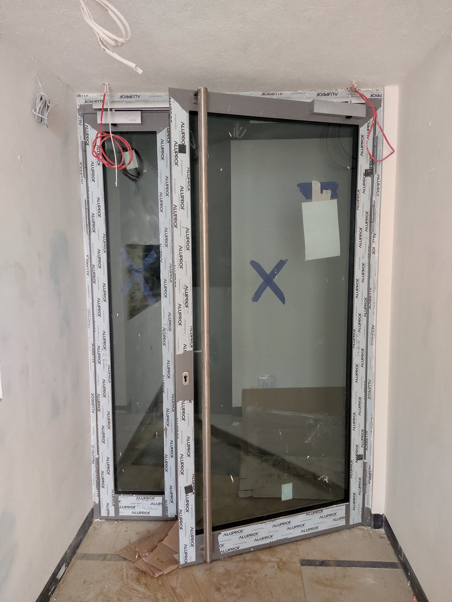 Montaż szklanych drzwi wahadłowych z aluminiową ramą, widoczne przewody instalacyjne i oznaczenia na szybie, sugerujące etap prac wykończeniowych.