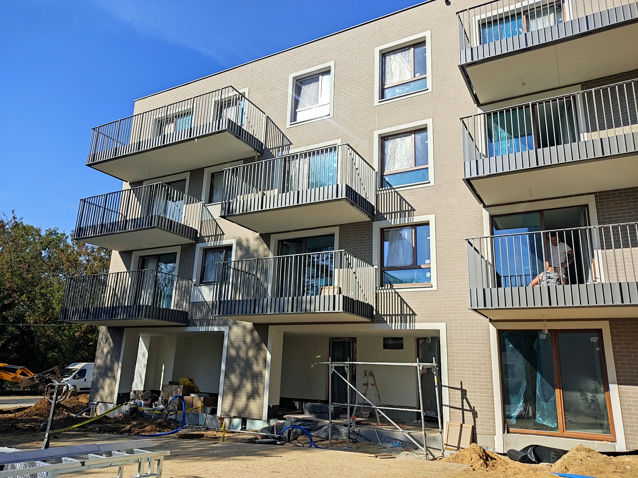 Nowoczesny budynek mieszkalny w trakcie budowy, widoczne okna z folią ochronną i balkony z metalowymi barierkami. Na dole rusztowania i materiały budowlane.