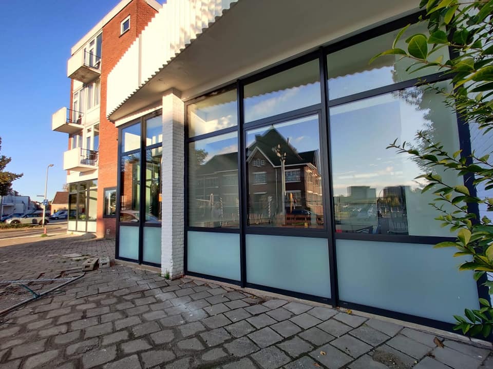 Elewacja budynku z dużymi oknami z czarnymi ramami i mleczną szybą u dołu, odbijającymi otoczenie. Widok z zewnątrz na chodnik z kostki brukowej.