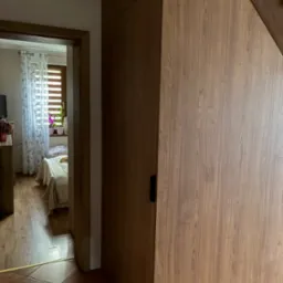 Drewniana zabudowa pod schodami z czarnym uchwytem, widok na pokój przez otwarte drzwi. Podłoga z terakoty.