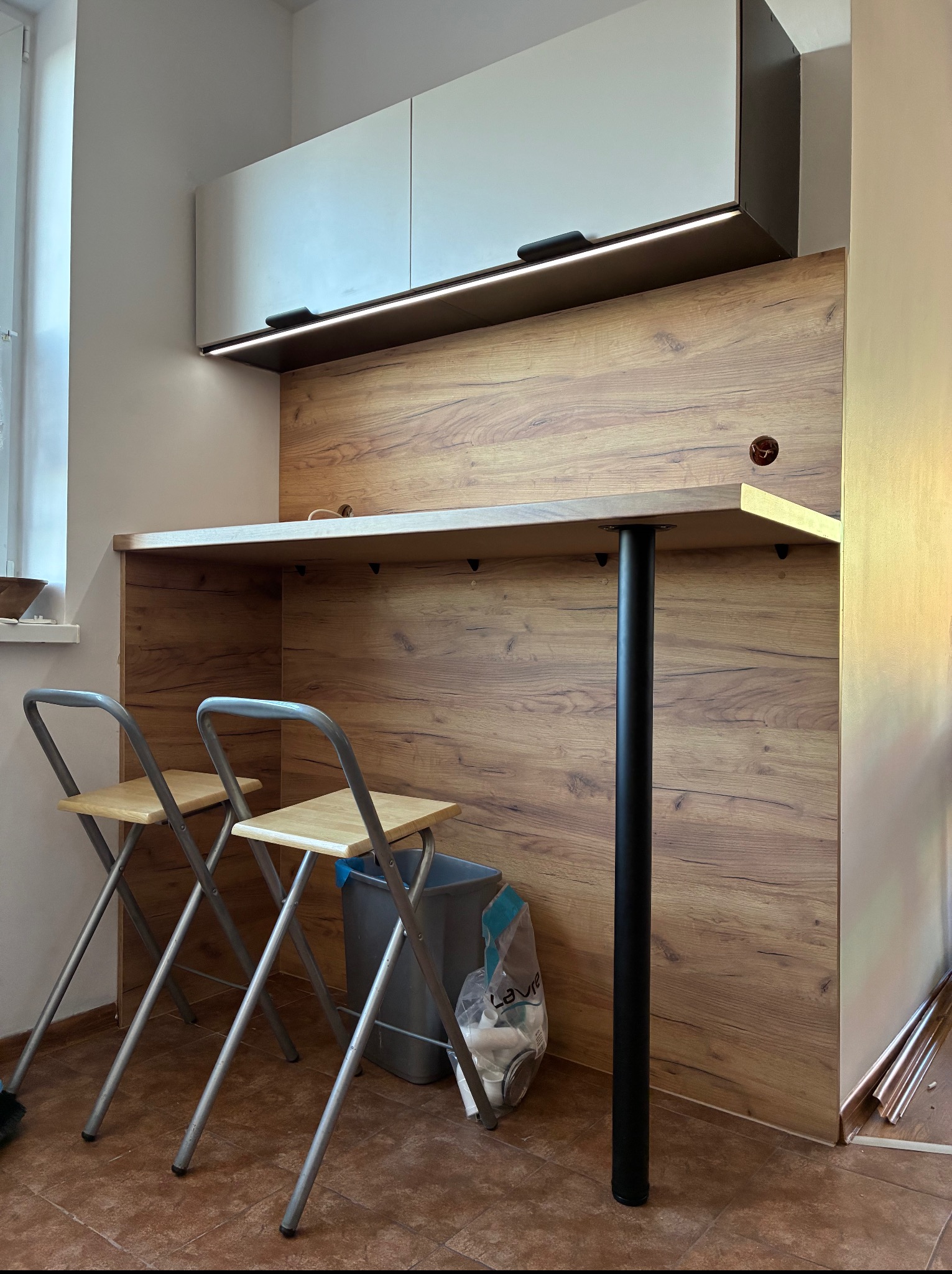 Drewniana zabudowa kuchenna z blatem i oświetleniem LED, uzupełniona o dwa składane krzesła barowe. Widoczny kosz na śmieci i fragment okna.