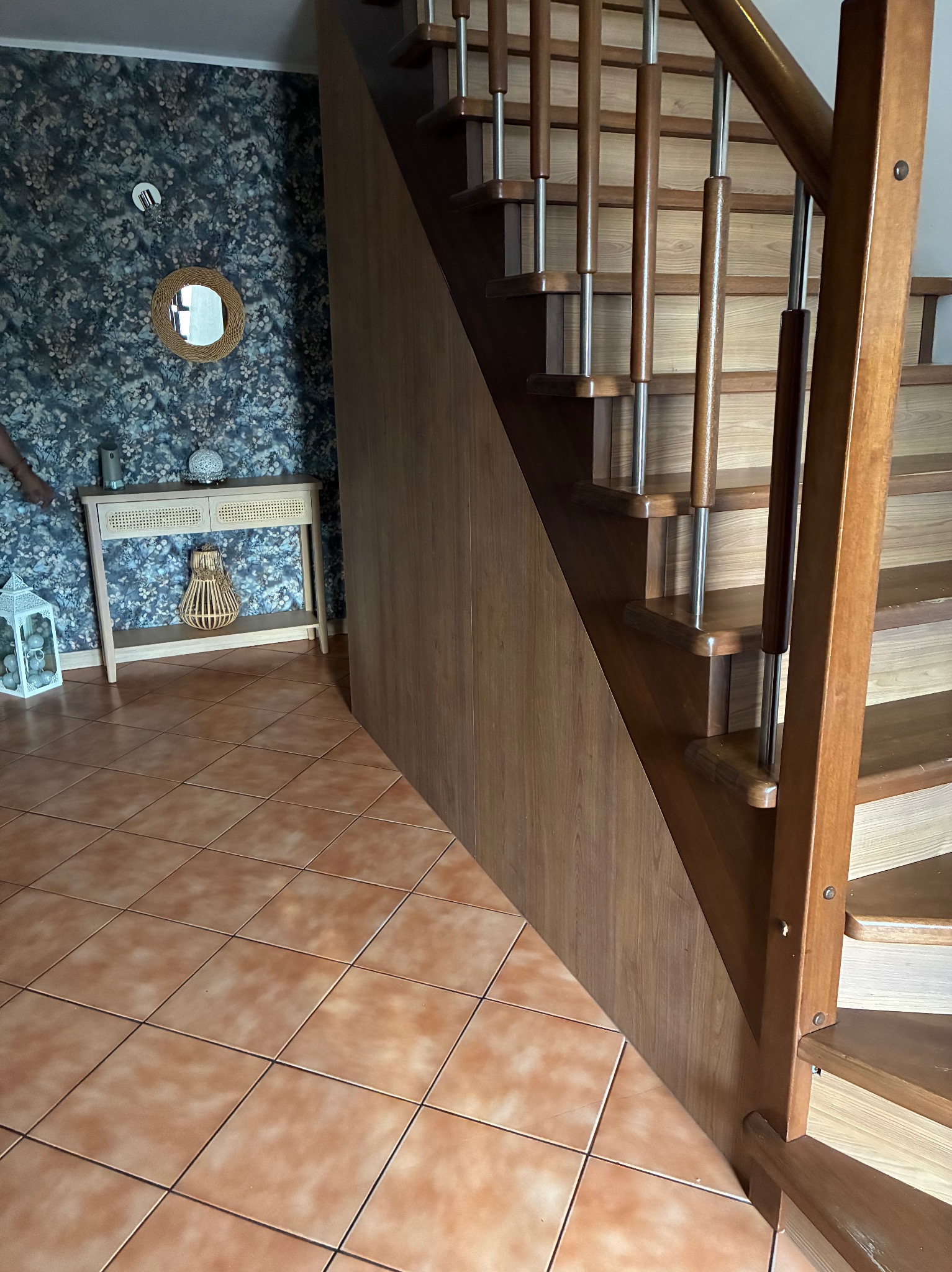 Drewniane schody z zabudową pod schodami w Kowalewie Pomorskim. Balustrady ze stali nierdzewnej. W tle konsola z lustrem i dekoracjami na tle wzorzystej tapety.