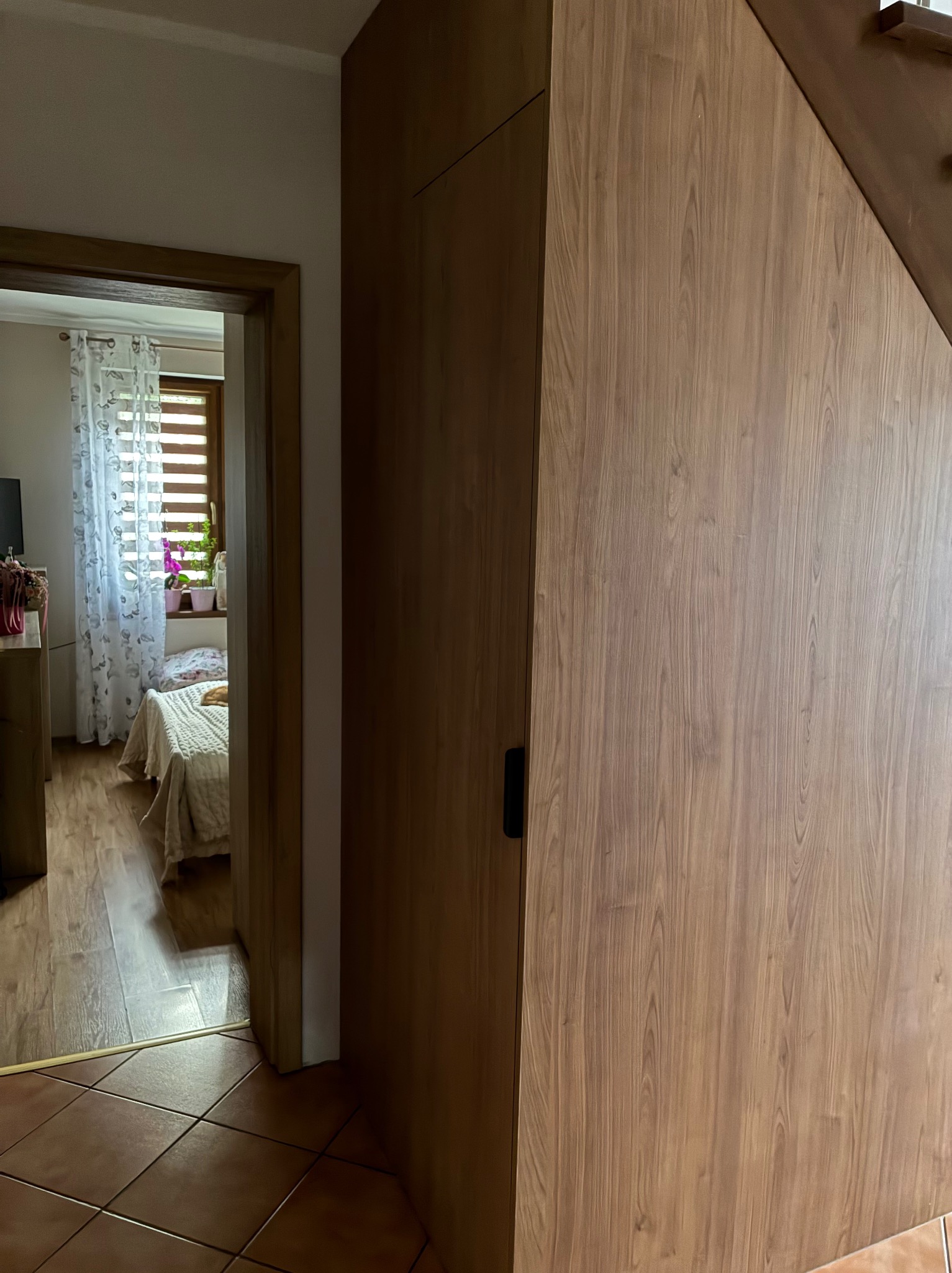 Drewniana zabudowa pod schodami z czarnym uchwytem, widok na pokój przez otwarte drzwi. Podłoga z terakoty.