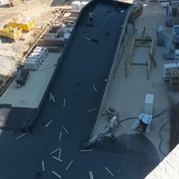 Dachy Plaskie Ewa Pupek - Prace dekarskie na dachu budynku w budowie. Widok z góry na czarną papę i dekarza z wałkiem. W tle budynki w stanie surowym, materiały budowlane i pomarańczowy silos.