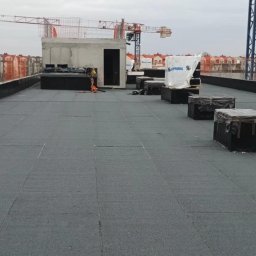 Dachy Plaskie Ewa Pupek - Rozległy, płaski dach pokryty czarną papą. W tle budynki w trakcie budowy, dźwigi i betonowa konstrukcja. Widoczne materiały budowlane i pracownik w oddali.