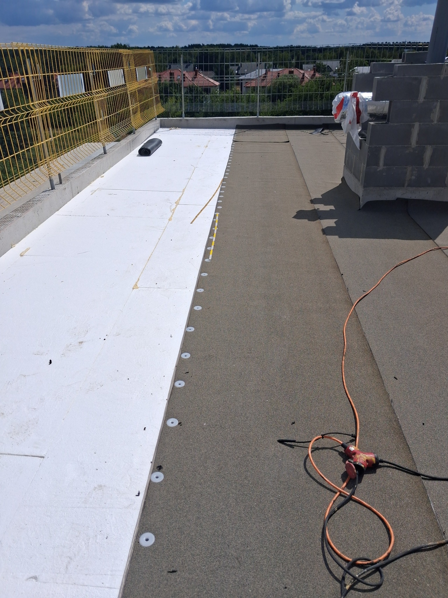 Prace dekarskie na dachu: izolacja termiczna z białego styropianu, mocowana mechanicznie do papy. Widoczne narzędzia i zabezpieczenia na krawędzi dachu.