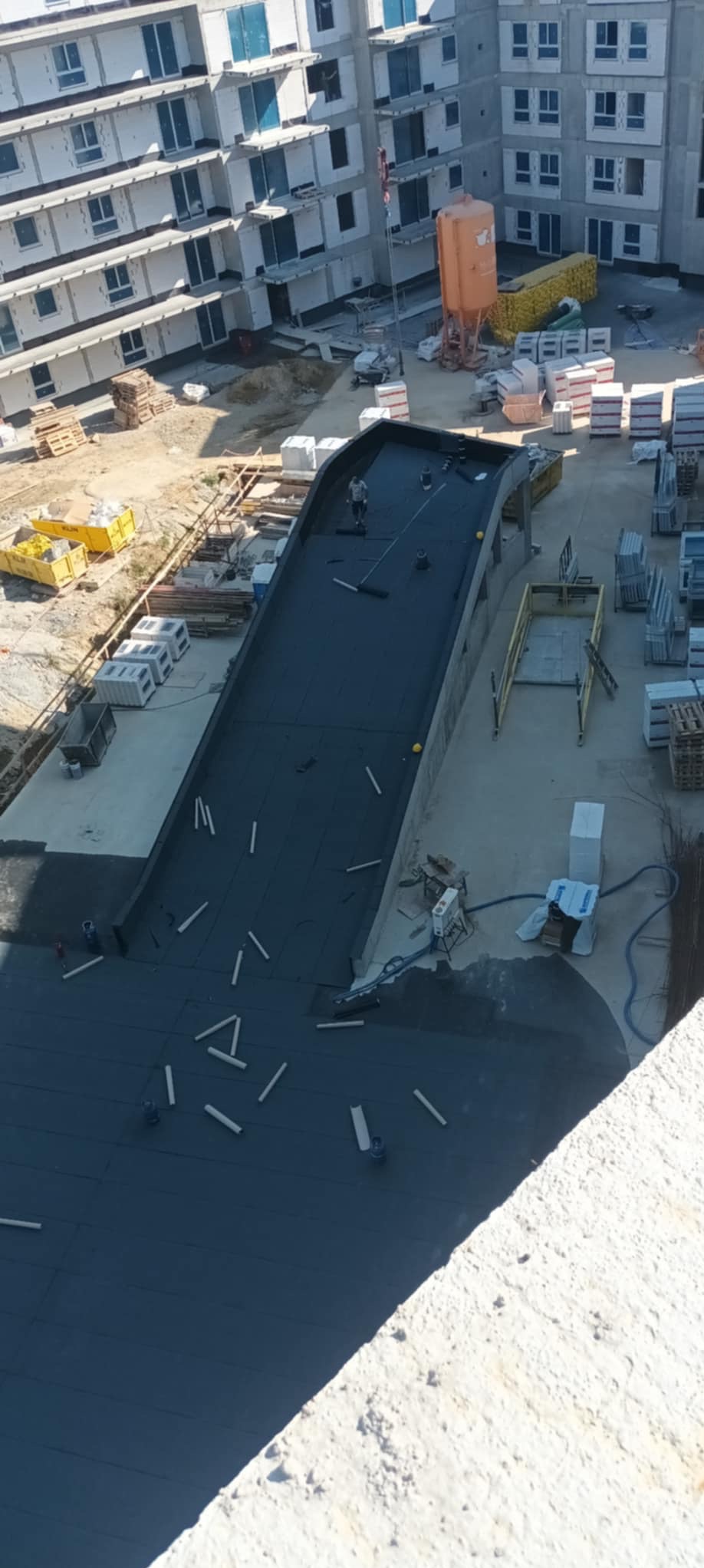 Prace dekarskie na dachu budynku w budowie. Widok z góry na czarną papę i dekarza z wałkiem. W tle budynki w stanie surowym, materiały budowlane i pomarańczowy silos.