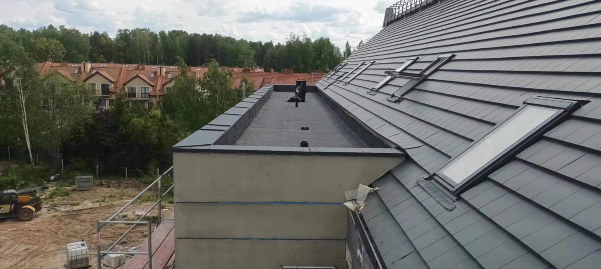Szary dach z dachówkami i oknami dachowymi, widok z góry na tle zieleni i zabudowy miejskiej. Fragment dachu płaskiego z papą. Prace dekarskie.