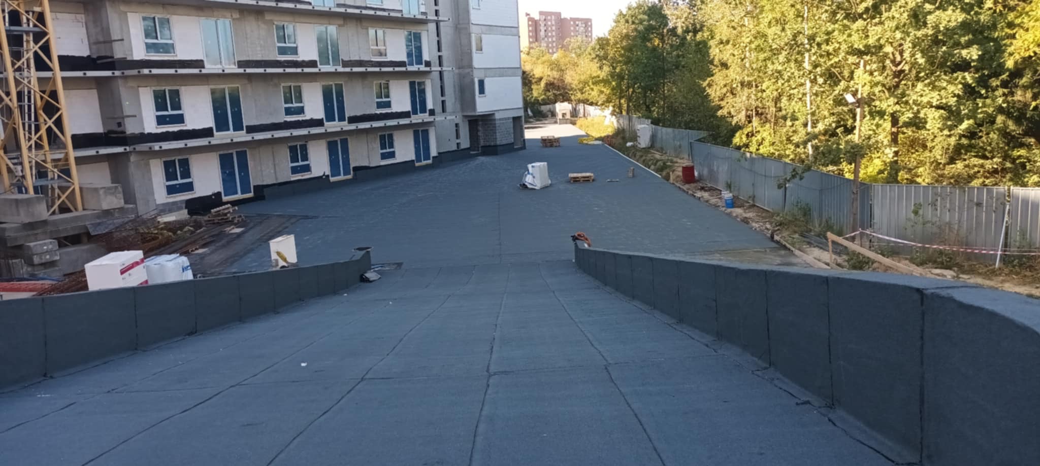 Wykończony dach budynku wielorodzinnego z papy termozgrzewalnej. Widoczne materiały budowlane, rusztowanie i fragmenty zieleni w tle.