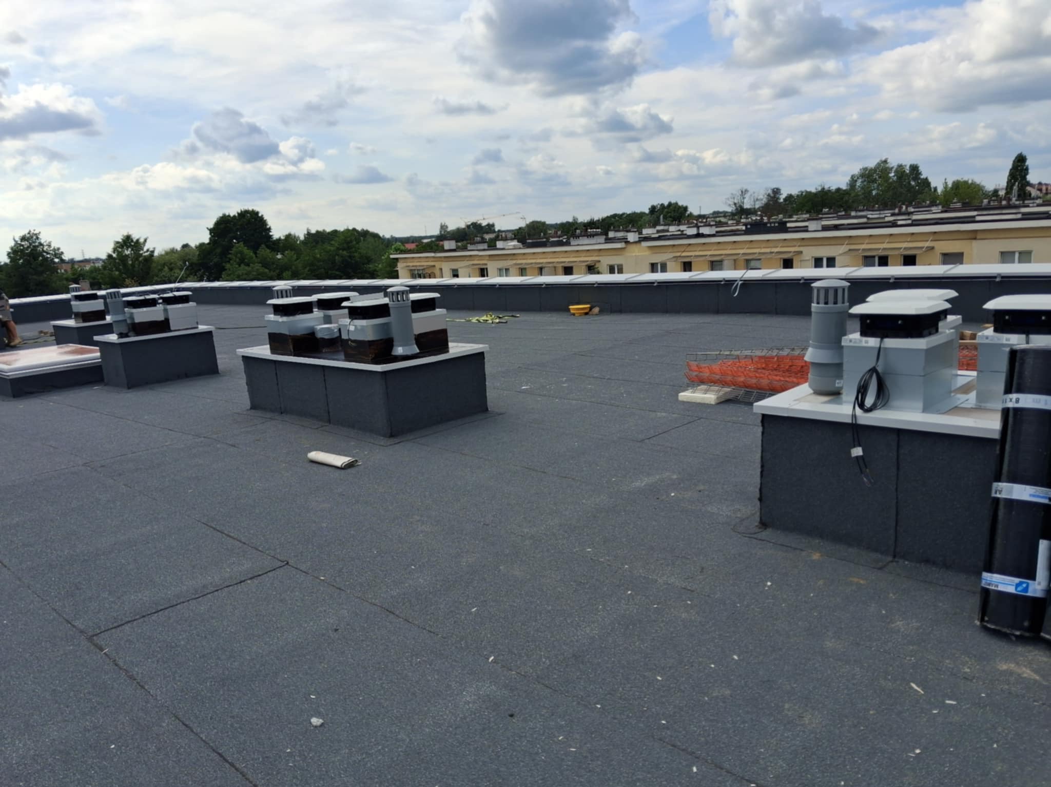 Widok na płaski dach z zamontowanymi systemami wentylacyjnymi i rolkami papy. W tle budynek i pochmurne niebo.