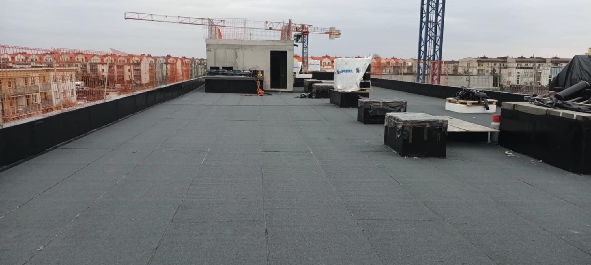 Rozległy, płaski dach pokryty czarną papą. W tle budynki w trakcie budowy, dźwigi i betonowa konstrukcja. Widoczne materiały budowlane i pracownik w oddali.